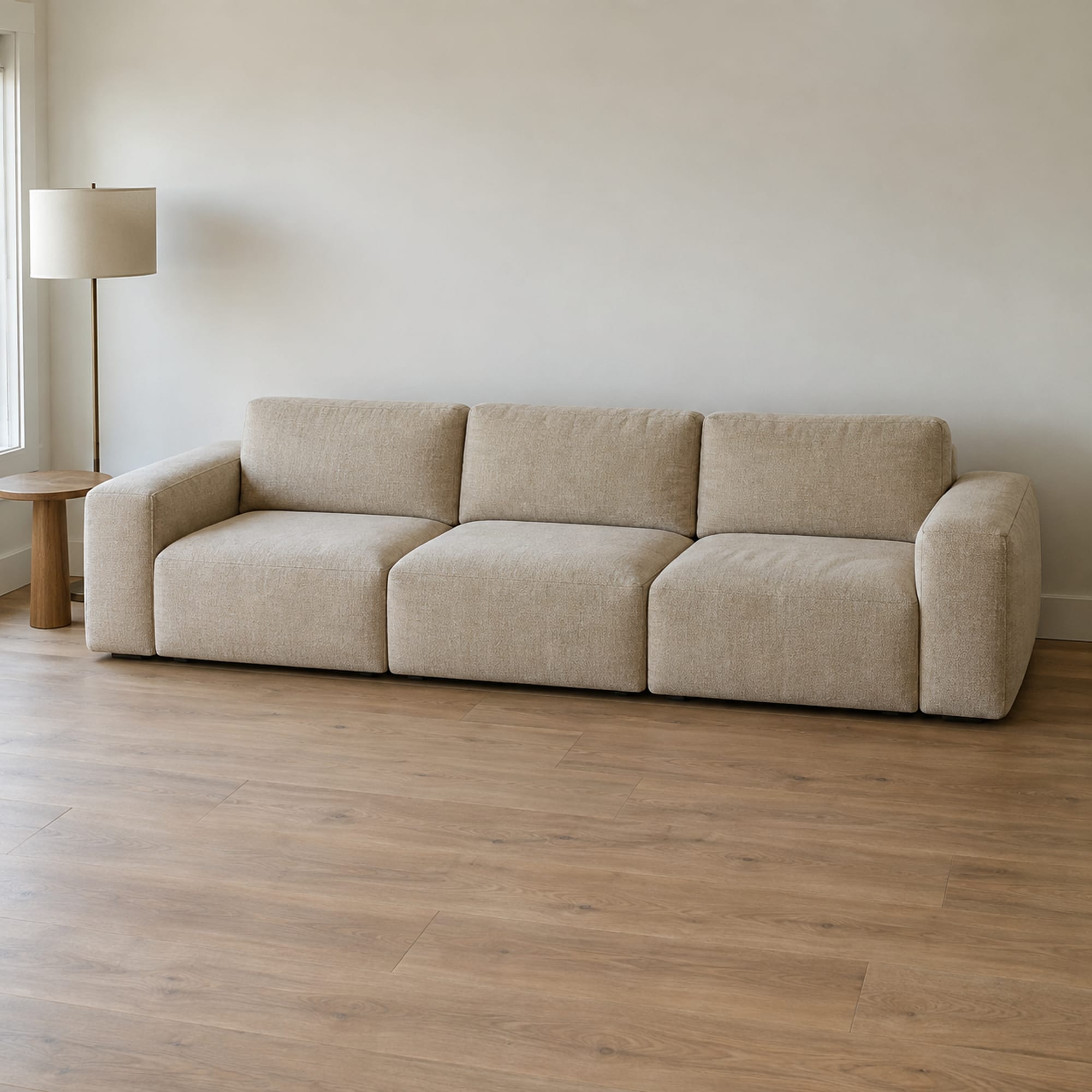 A three-seat modular sofa of separate upholstered seat blocks, low back pieces and arm pieces in a warm sand chenille, set against the long wall of a modestly furnished daylit living room with a small round side table and a plain floor lamp at one end, photographed from a raised front three-quarter angle at standing height so the whole piece and the room around it read together and the lines between the blocks show only as soft seams