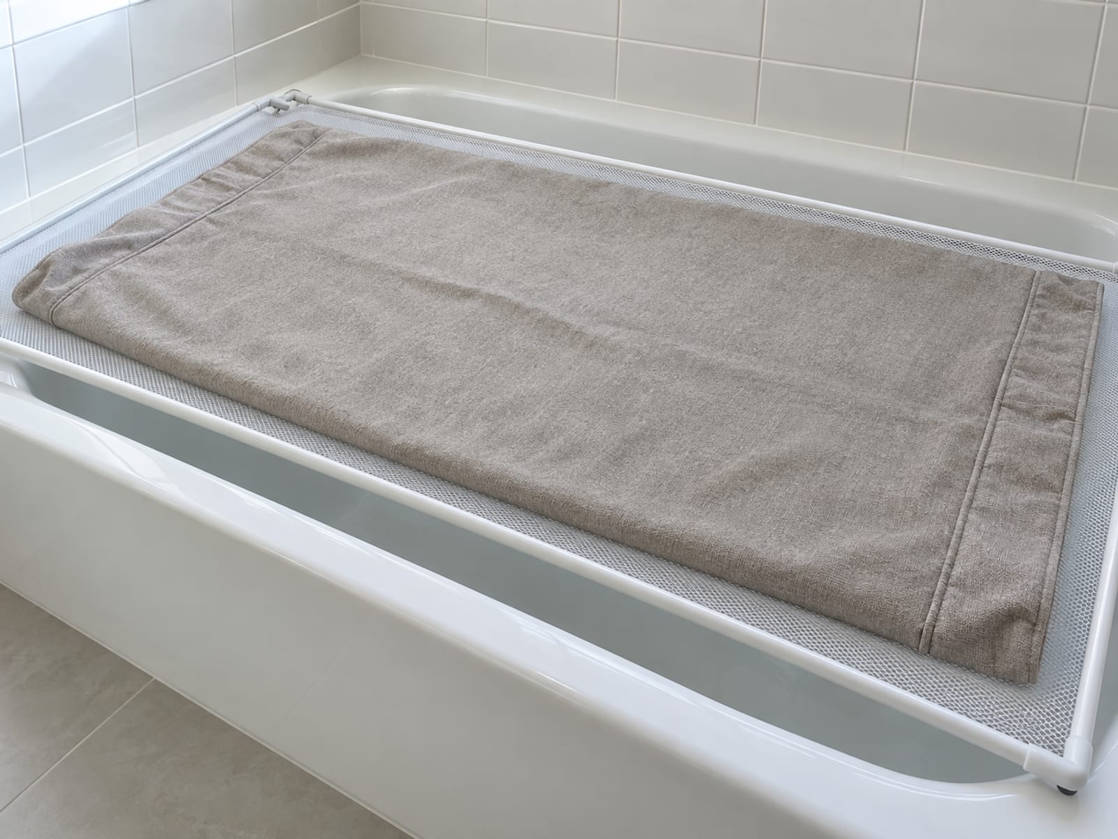 One removed seat cover from a modular sofa laid out flat and smooth on a plain white mesh drying screen resting across the rim of an ordinary white bathtub in a plain tiled bathroom, photographed from standing height at a slight downward angle