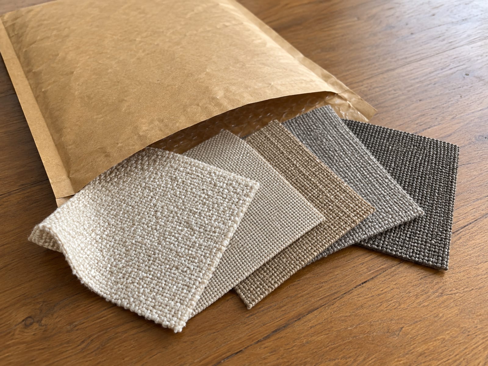 A plain unmarked padded paper mailer lying on a bare wooden hall table with its flap open and five small square fabric samples in five different weaves in the warm neutral range sliding part way out in a loose overlapping fan, photographed from standing height at a slight downward angle