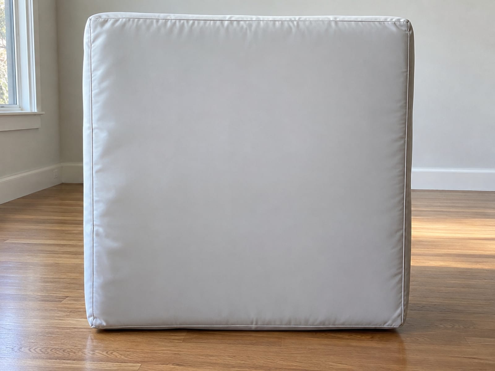 One square foam seat insert from a modular sofa standing upright on its edge on bare oak floorboards with its fabric cover removed, fully enclosed in a smooth pale waterproof liner with neatly sealed edges, photographed square on from low standing height