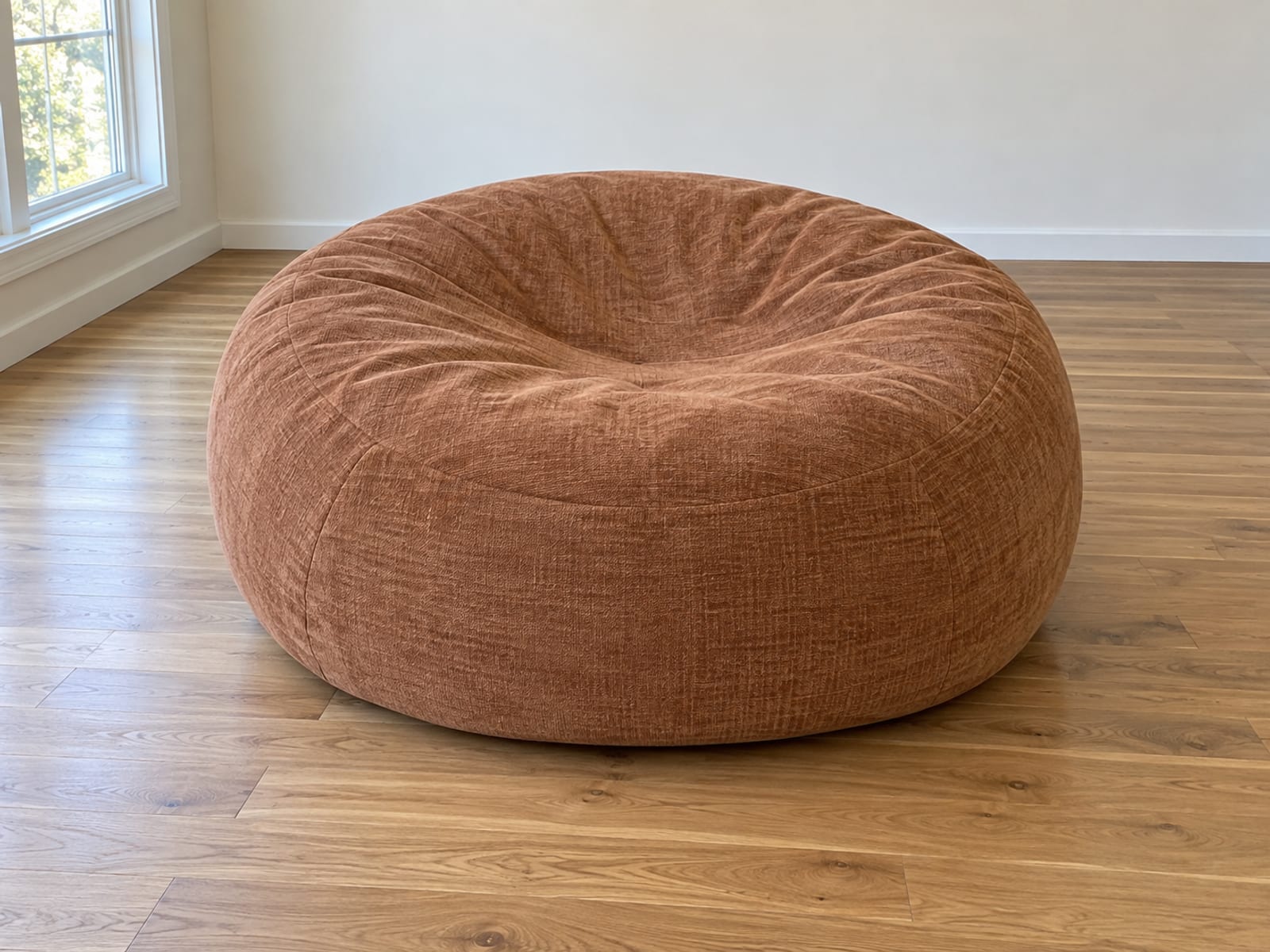 One very large round foam-filled floor lounger in a short dense chenille cover in a warm clay tone resting on bare oak floorboards in an empty daylit room, photographed from standing height at a slight downward angle