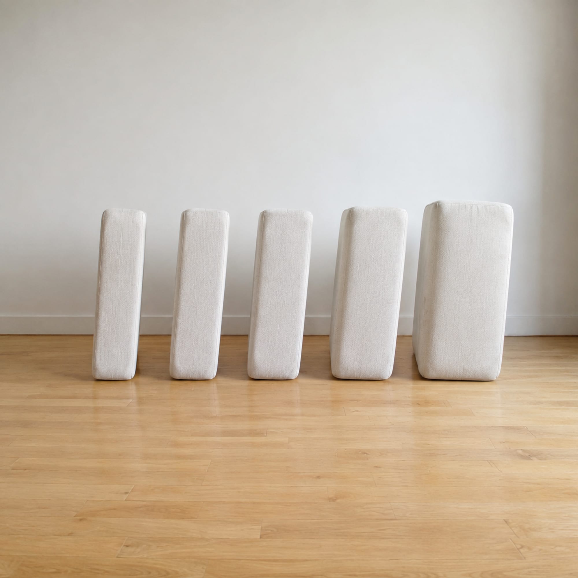 Five plain pale seat cushions of clearly different front-to-back depth standing upright in a row on bare floorboards, shallowest on the left and deepest on the right