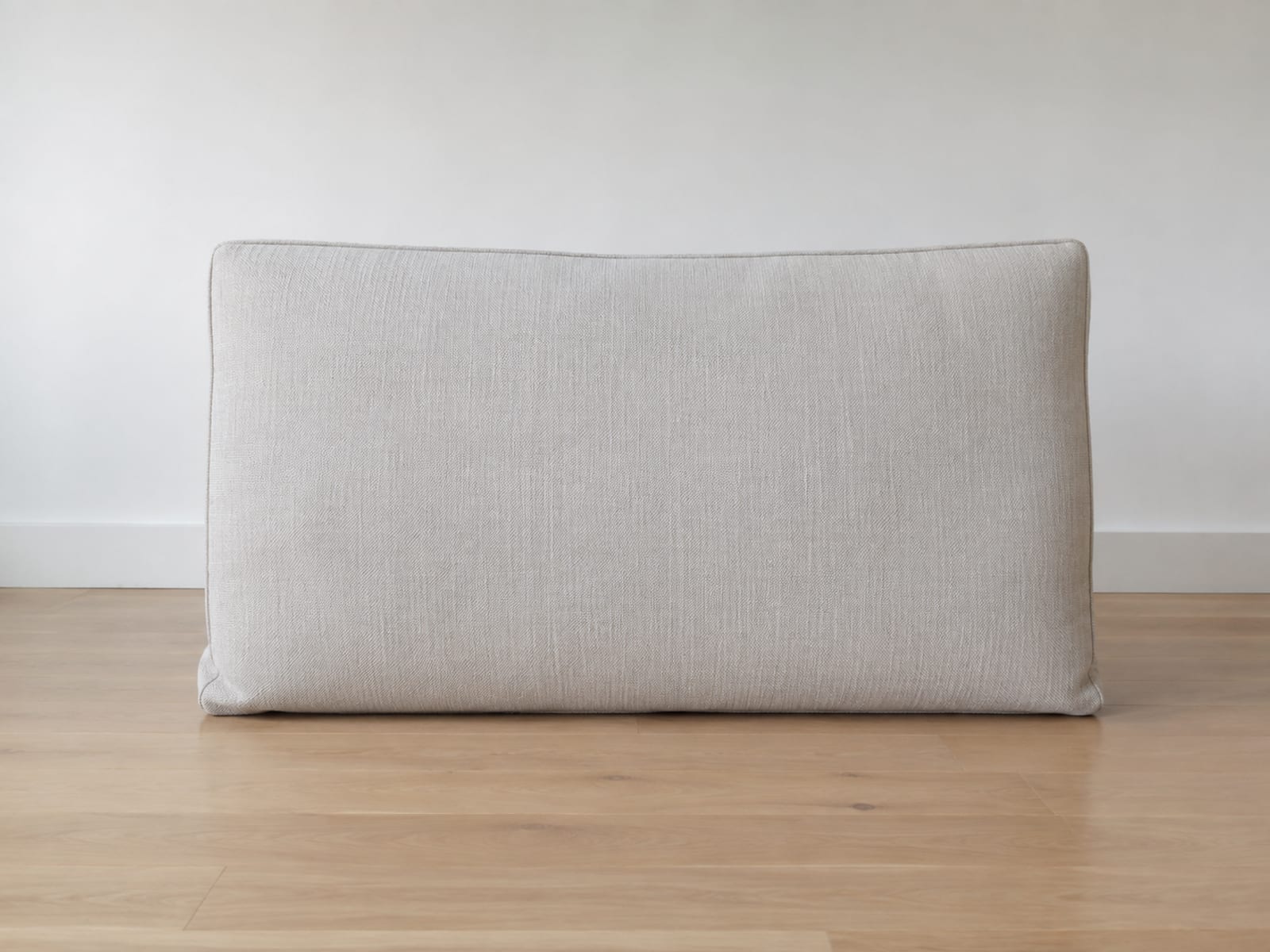 One large plain pale back cushion standing upright on its long edge by itself on bare floorboards, leaning very slightly, with clear floor either side