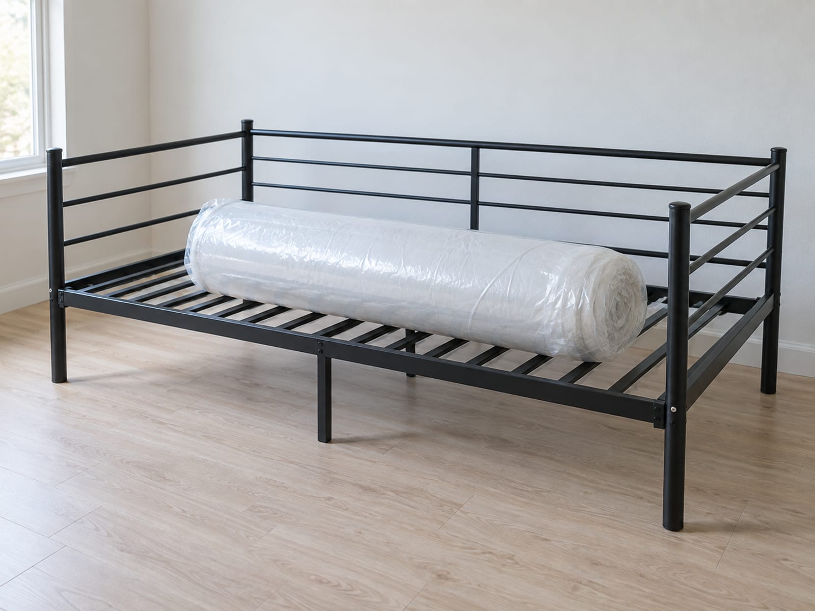 A newly assembled plain metal daybed frame with no mattress in it, and a separately bought twin mattress still tightly rolled in plain clear wrap lying across the bare slats of the frame, photographed at a front three-quarter angle