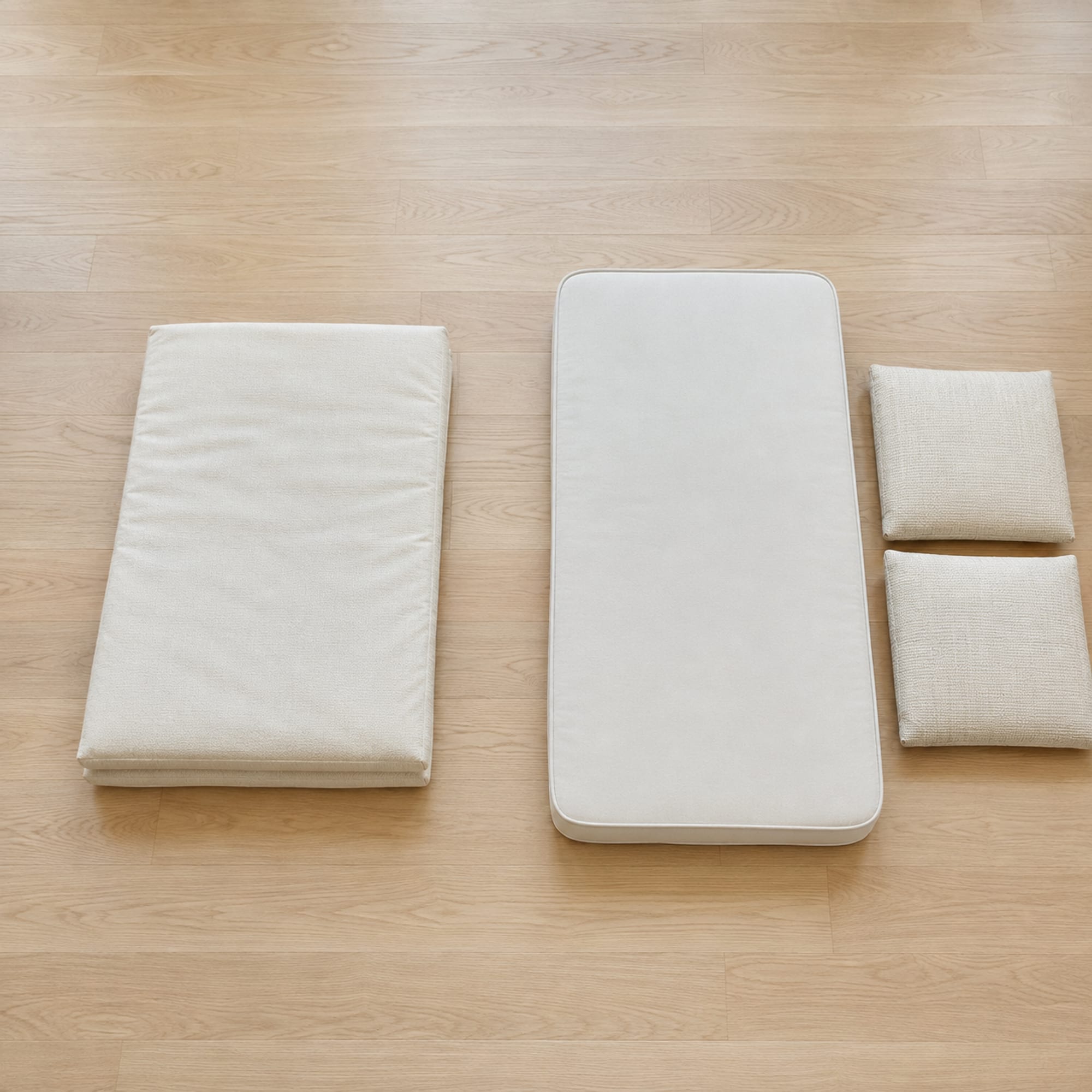 Two groups laid out side by side on bare oak floorboards seen from above, on the left a single folded futon pad and on the right a thin bare mattress with two separate seat cushions stacked beside it