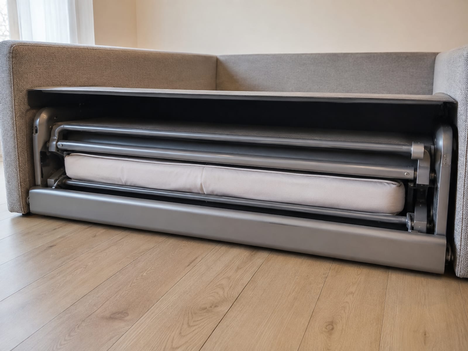 A low close view into the open front of a sofa bed frame with its seat cushions removed, showing the folded steel mechanism and the edge of the thin mattress packed inside
