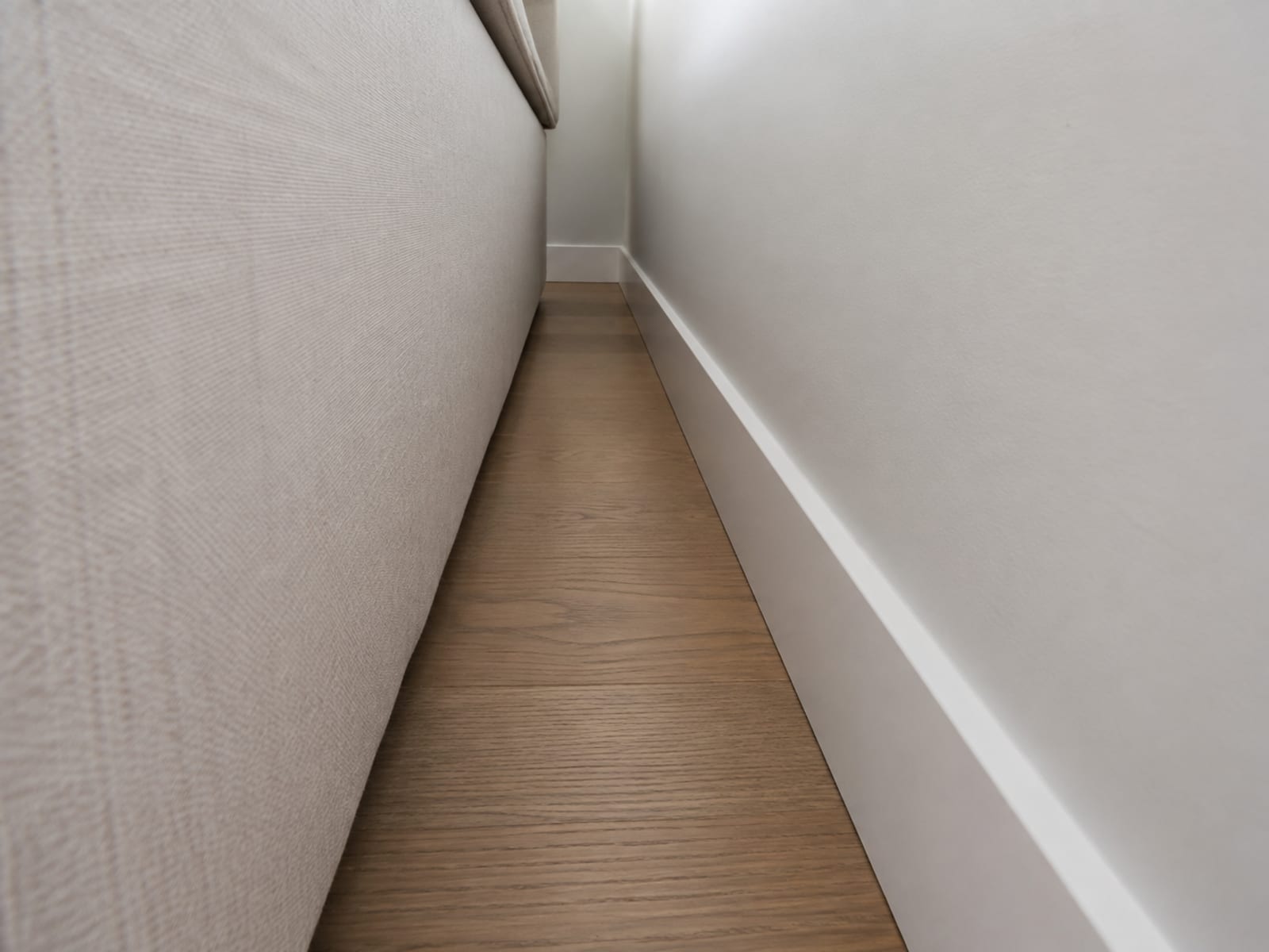 A low view along the narrow gap between the back of a pale sofa and a plain painted wall, the baseboard running along the bottom of it