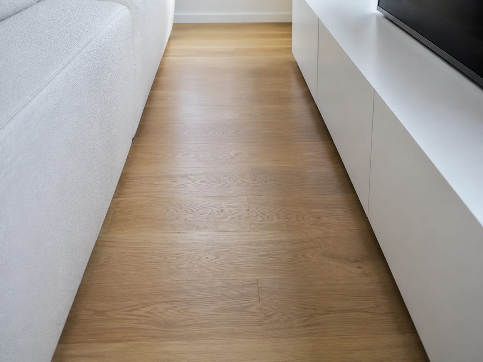 A narrow strip of bare floorboards running between the front edge of a sofa and the base of a low television console, seen down its length