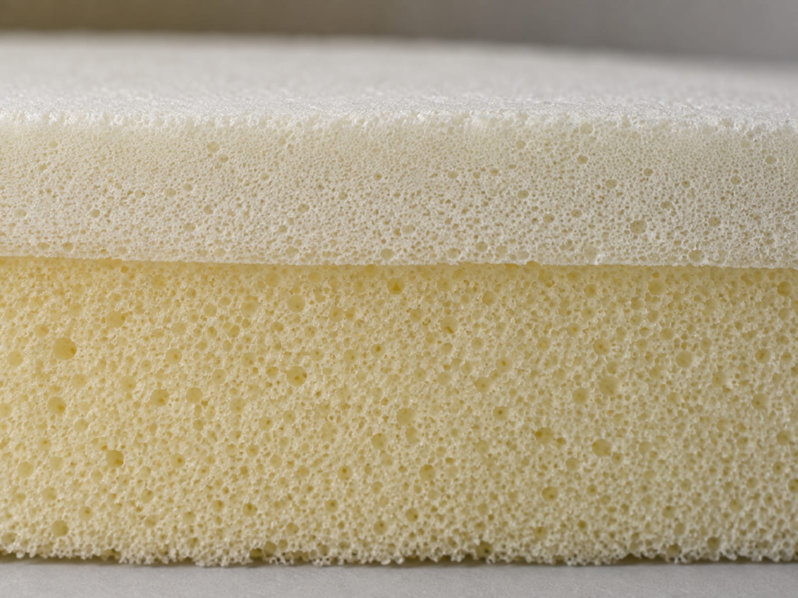 An extremely close view of the clean cut face of a seat cushion core showing two bonded layers of foam, a thinner pale memory foam layer on top of a thicker firmer base layer, the fine open cell texture of each plainly different under soft raking light