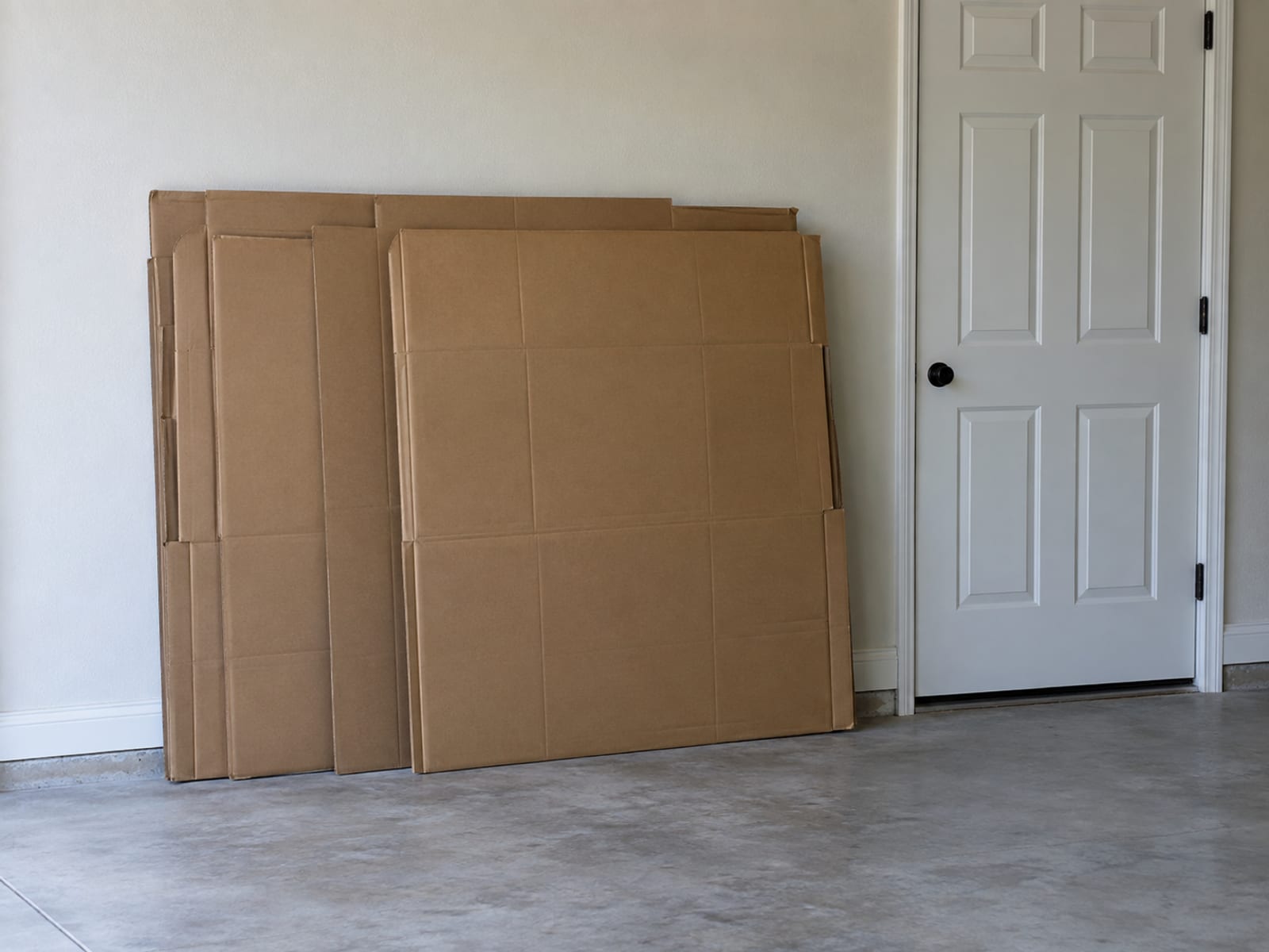 The sofa's original cartons flattened and kept, standing neatly on their long edges against the plain wall of a clean empty garage beside a closed interior door, photographed square on from standing height