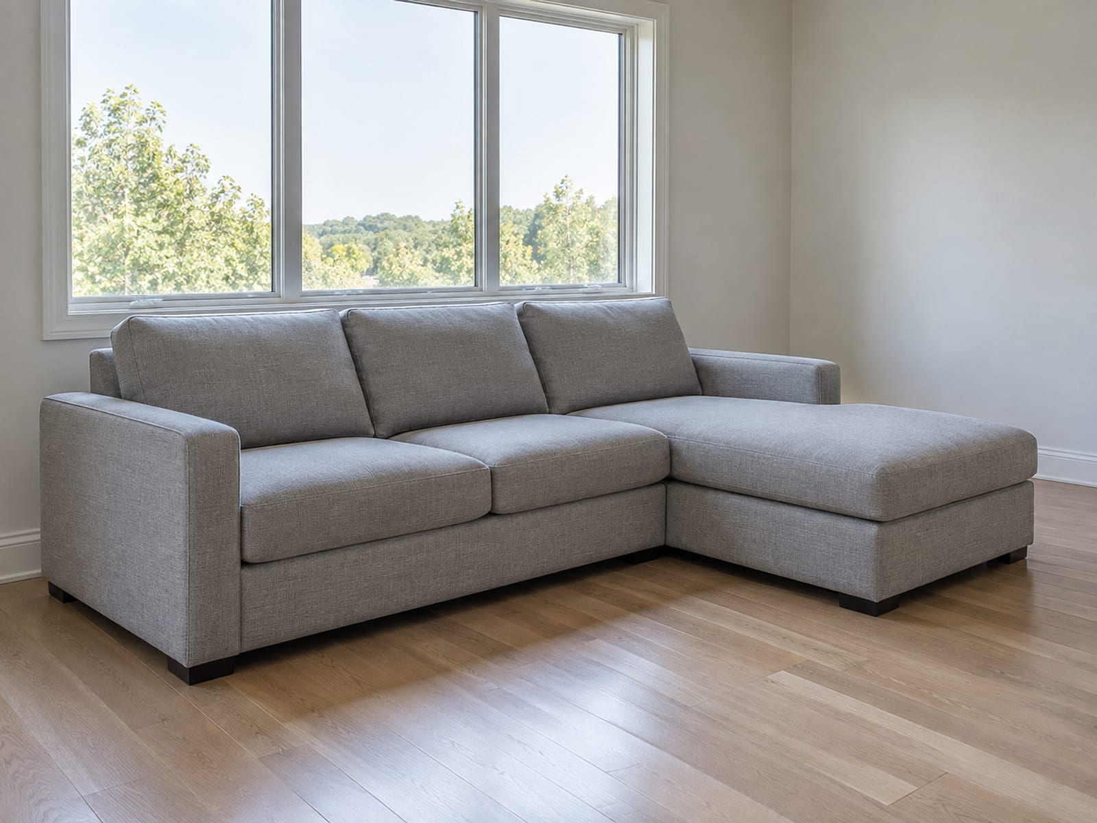 A compact sofa-with-chaise sectional in a pale gray weave standing beneath a wide window in a small daylit apartment living room, its chaise on the far side, photographed from the near end of the room at standing height at a front three-quarter angle