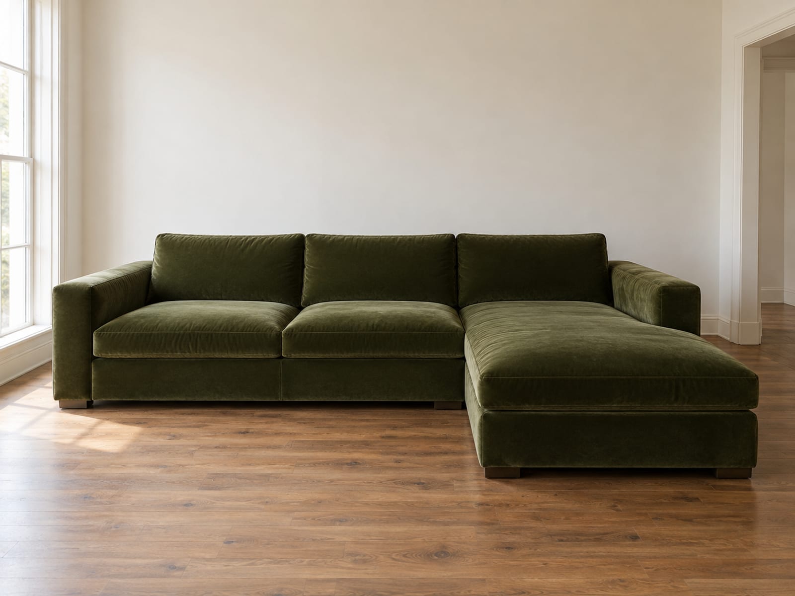 A sectional upholstered in a rich deep olive cotton velvet standing in a daylit living room with warm white walls and bare floorboards, photographed from the front at standing height a little off center