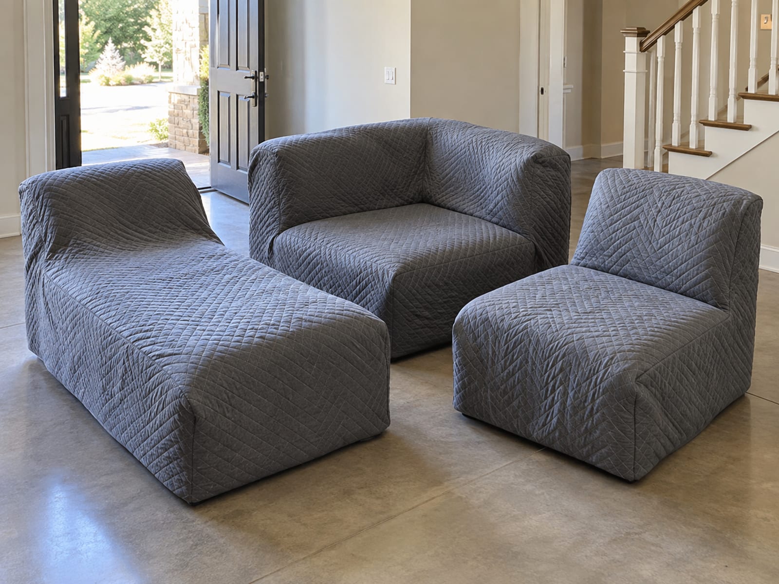 The separate pieces of a sectional just carried in, each fully wrapped in plain gray quilted moving blankets and standing in a neat group on the floor of a daylit entry hall beside the open front door, their shapes readable through the blankets as a corner unit, a chaise and an armless seat