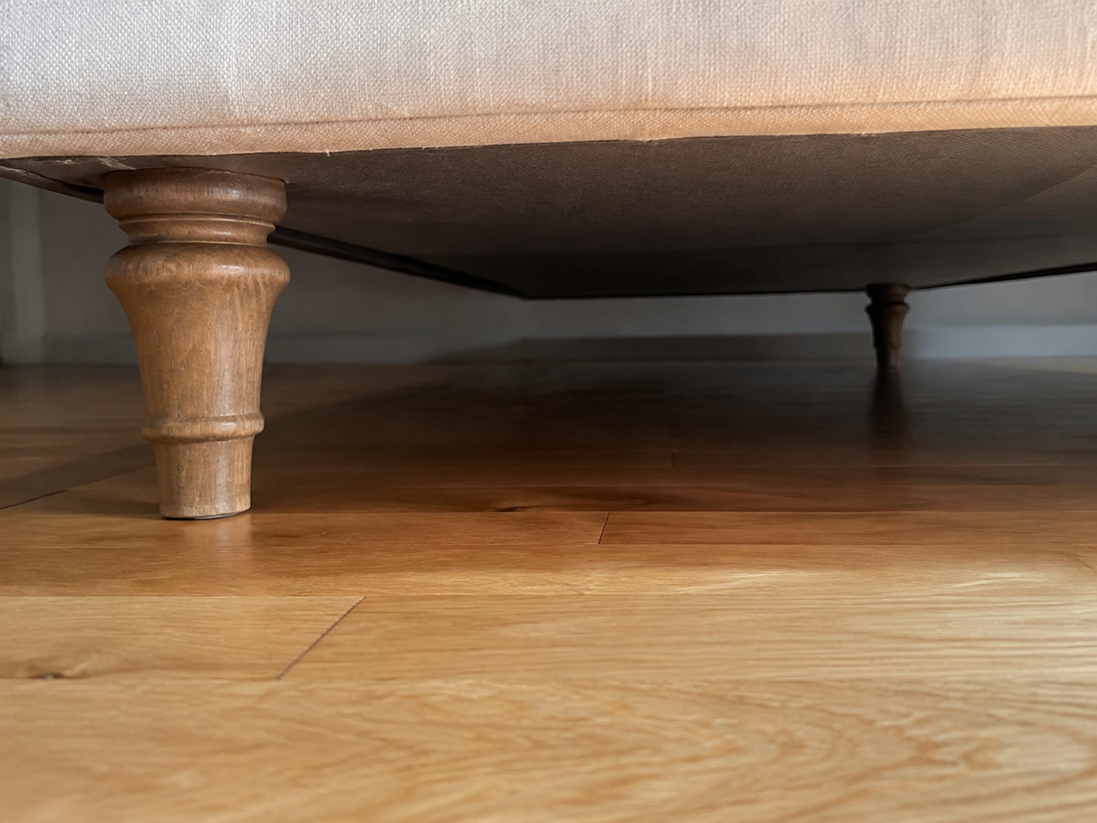 A low view into the shadowed space under a sofa, the front legs and the underside of the frame above clean bare floorboards running back into darkness