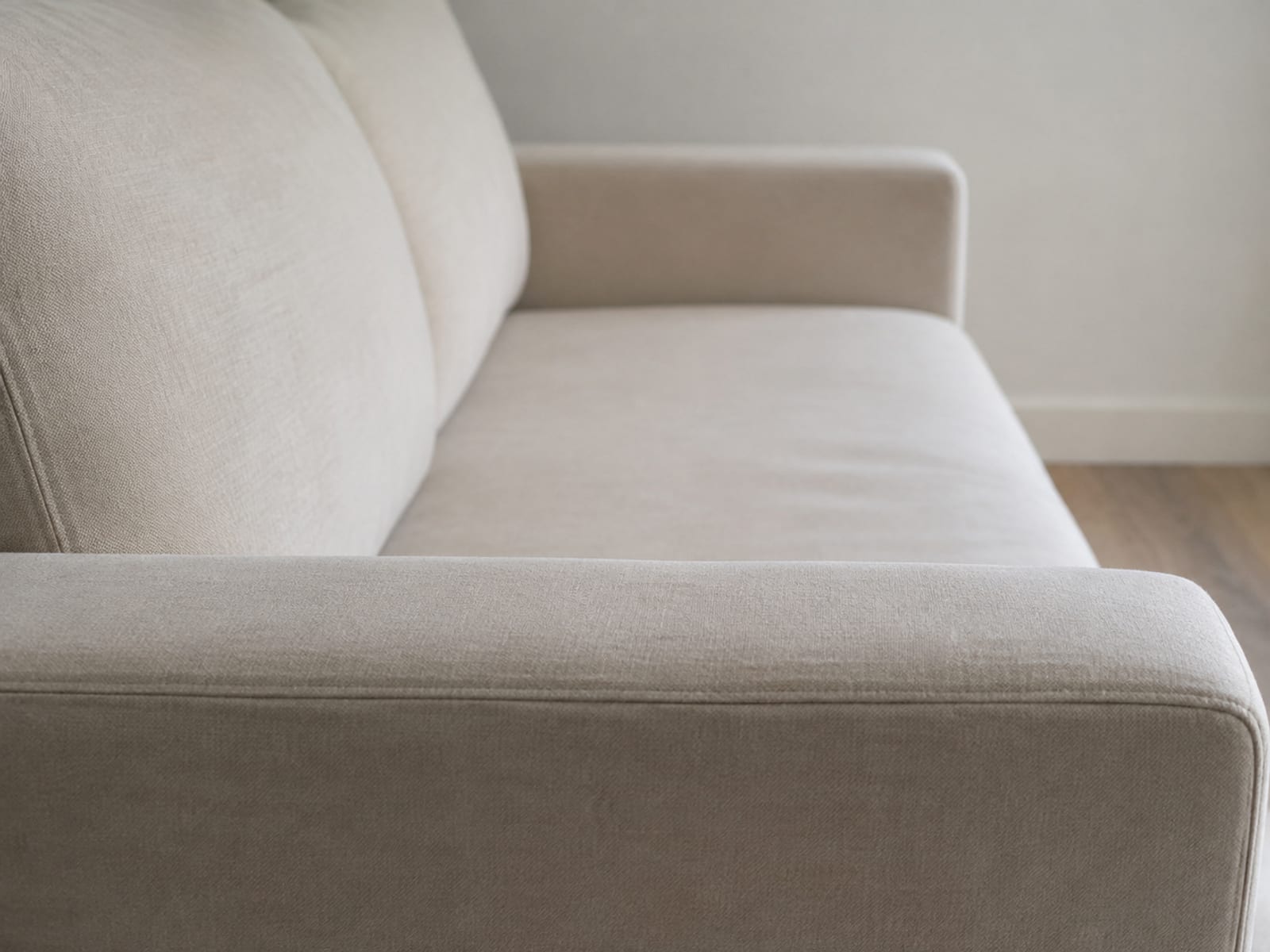 Three fabric swatches in three warm neutral tones laid flat on bare oak floorboards against the foot of a painted baseboard, seen from a low angle with the wall above