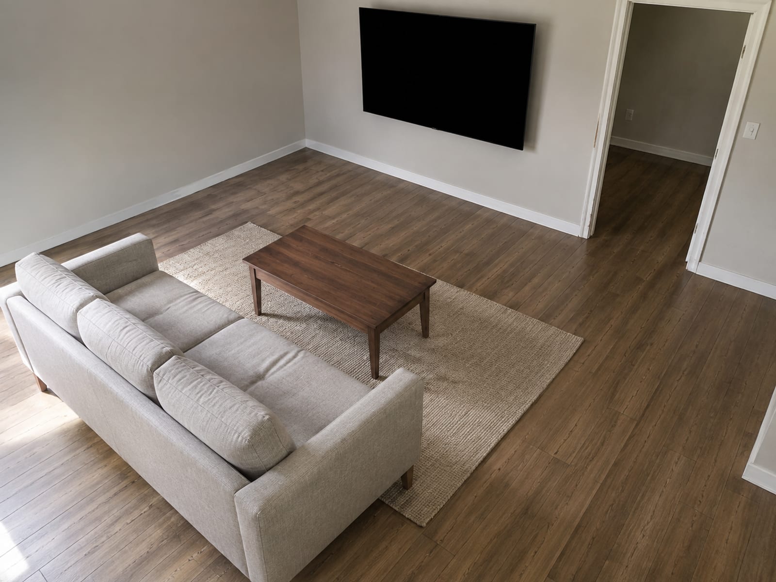 A small living room seen from a high corner, sofa facing a switched-off wall-mounted screen, with open walking room around it