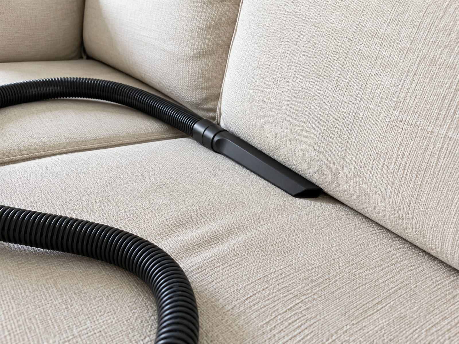 The narrow crevice tool of a domestic vacuum resting in the open gap between a seat cushion and the sofa back, its hose lying slack across the seat
