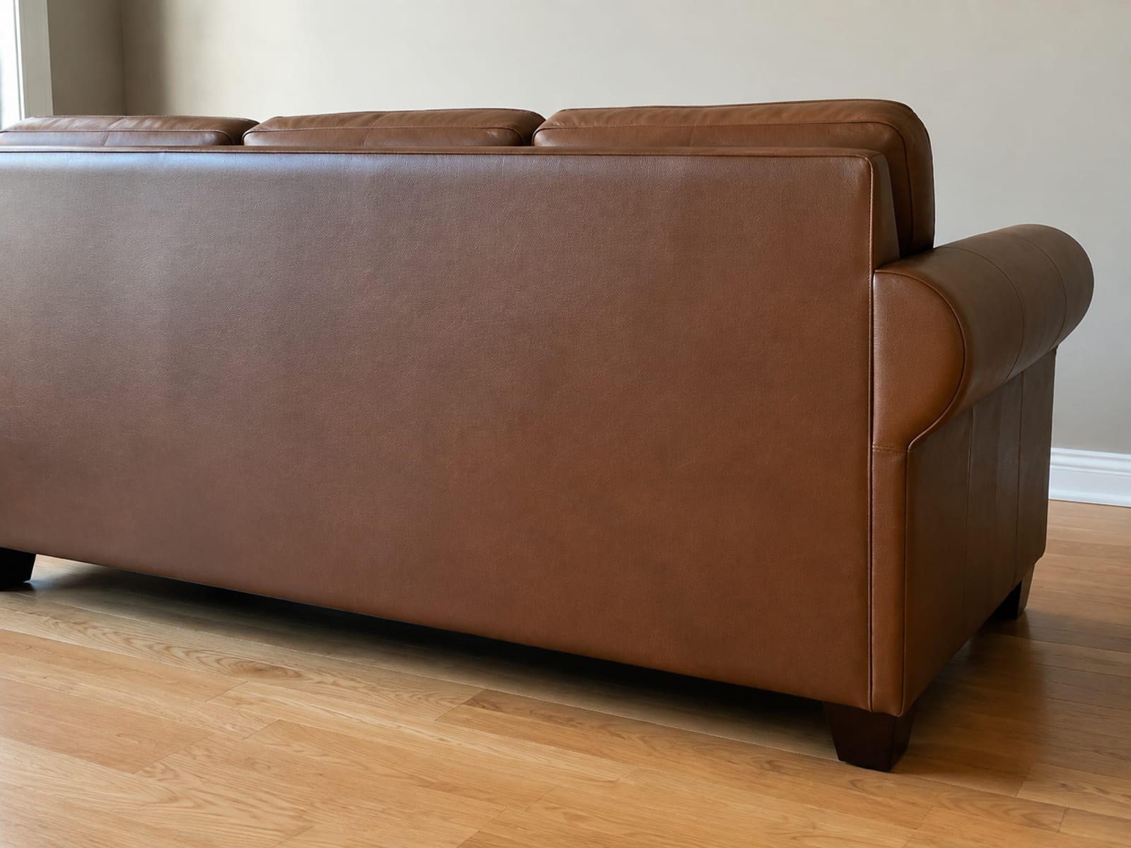 A leather three-seat sofa seen from behind, standing clear of a plain wall, its outside back panel completely even and unworn