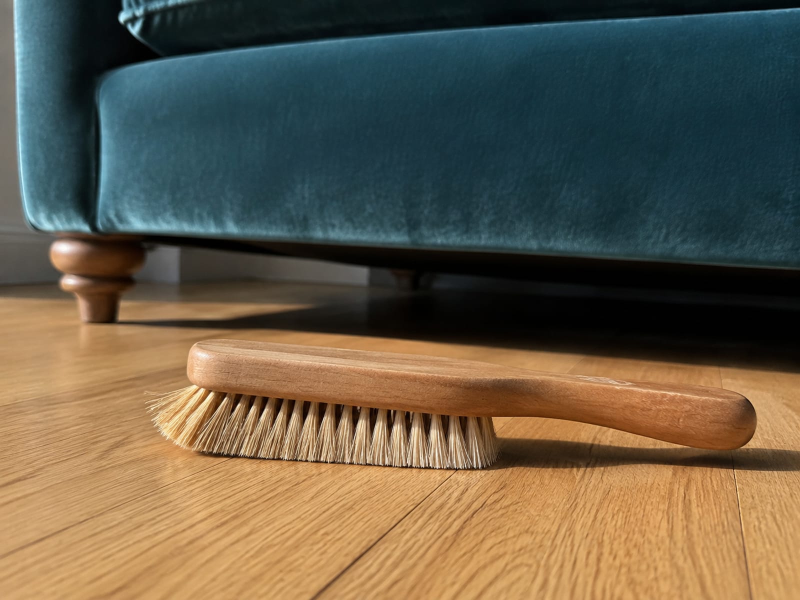 A wooden-handled brush with pale natural bristles lying on its side on bare floorboards beside the foot of a velvet sofa