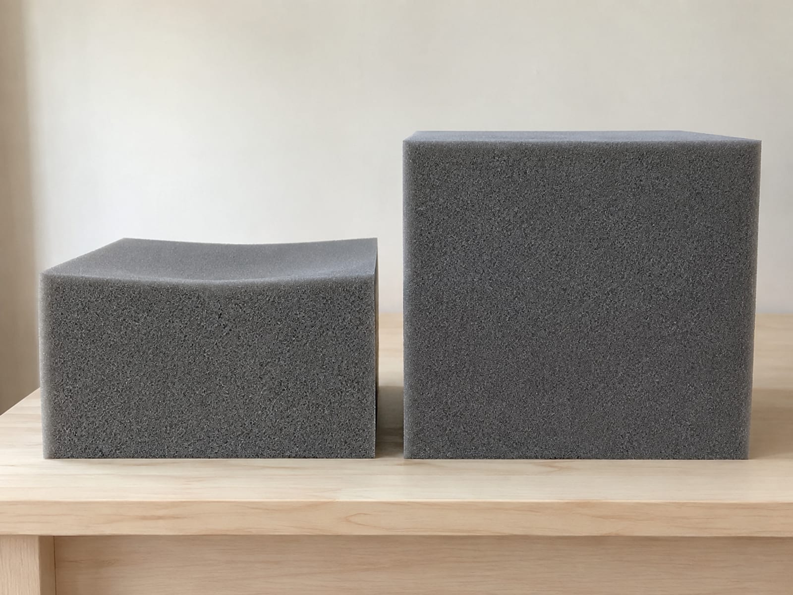 Two plain gray foam blocks of the same footprint standing side by side on a table, the left one shorter with a settled hollow top and the right at full square thickness
