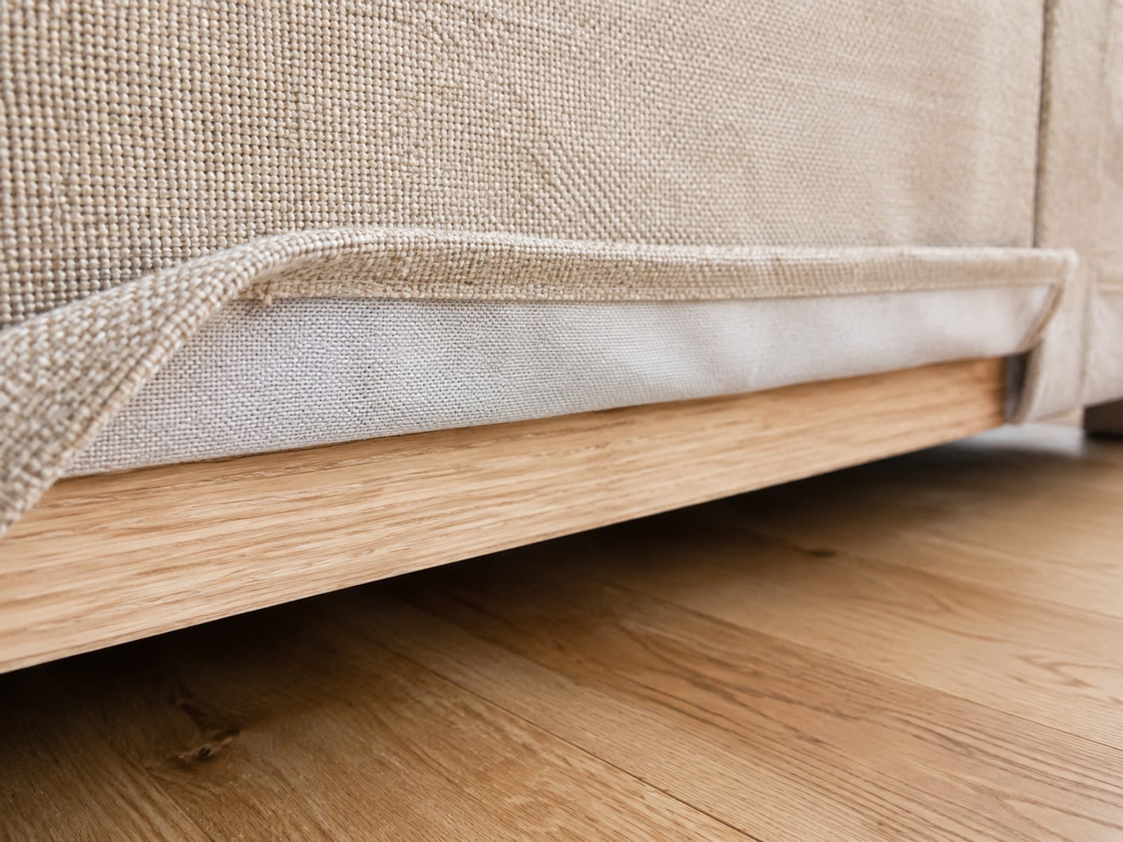 The lower hem of a sofa skirt turned up and tucked over the frame rail, showing the concealed reverse face of the fabric