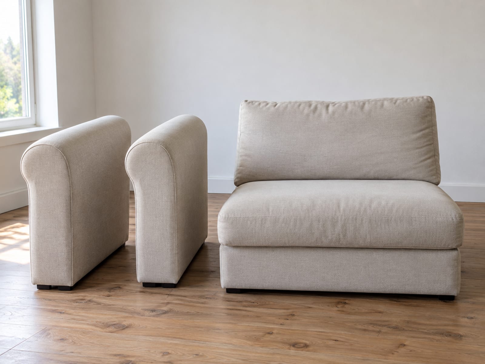 A sofa whose two bolt-on upholstered arms have been detached and stand upright side by side on bare floorboards next to the armless seat and back in an empty daylit room, photographed from standing height at a front three-quarter angle