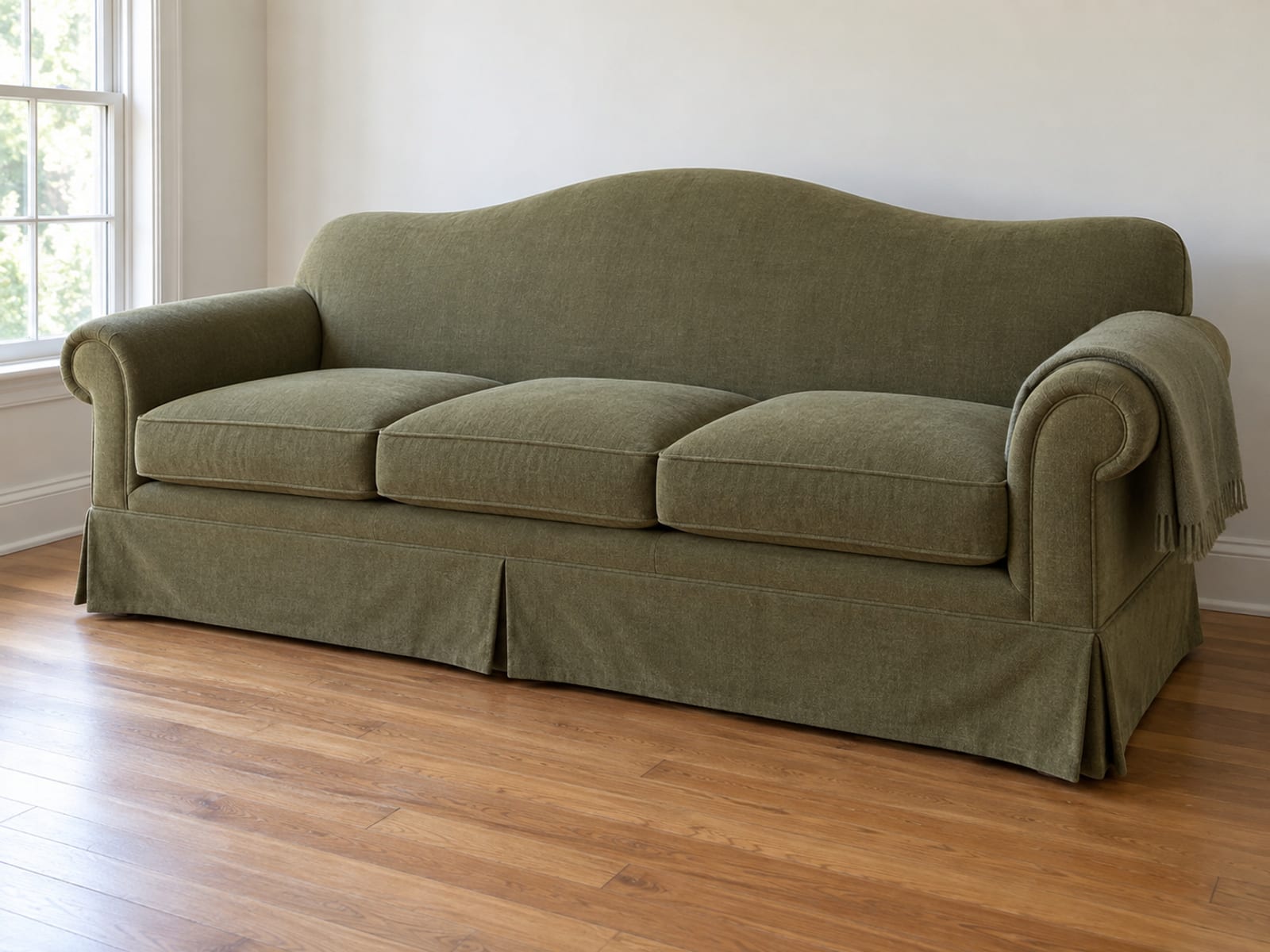 The same kind of older camelback sofa in plain moss green standing against a plain light wall beside a window, its cushions plumped and squared and one plain folded throw laid over one arm, on clean bare floorboards in bright even daylight, photographed from a front three-quarter angle at standing height