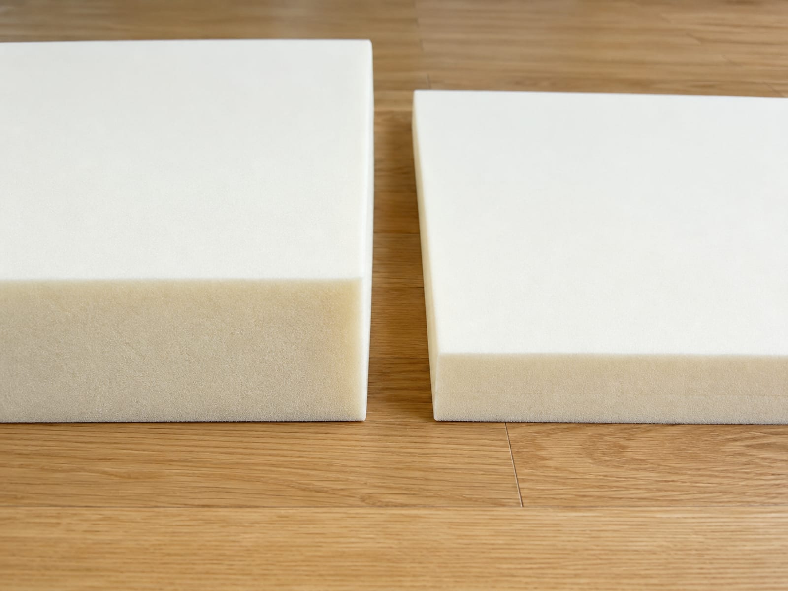 Two plain foam toppers of clearly different thickness lying flat side by side on bare floorboards with a gap between them, both cut edges facing the camera