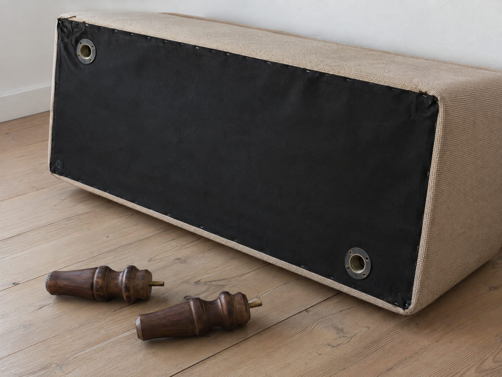 A sofa tipped onto its back on a bare floor with two wooden legs unscrewed and lying beside it, the empty mounting plates visible underneath