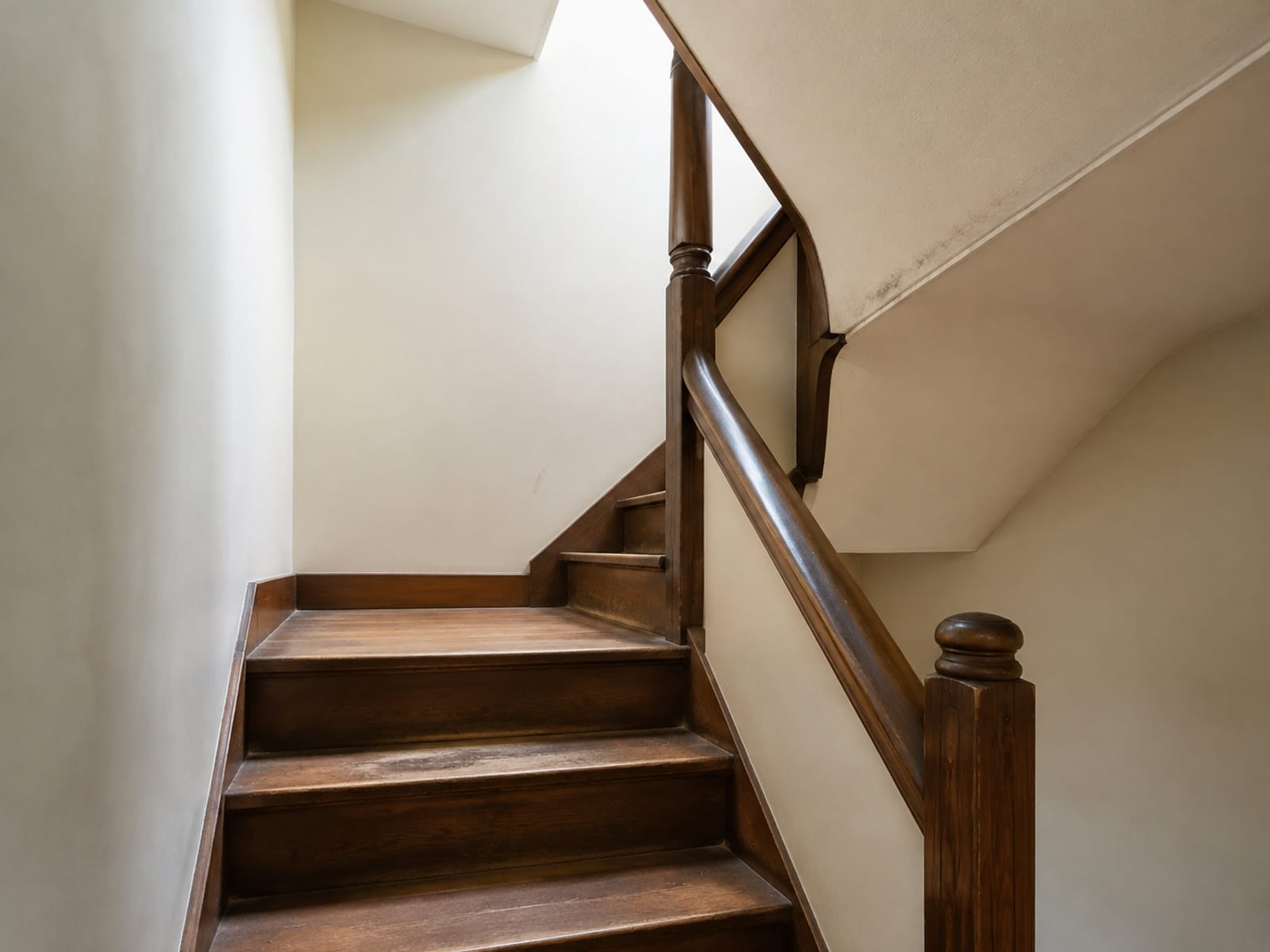The half-landing of a staircase that turns back on itself, seen from below, with the sloping ceiling coming down toward the handrail