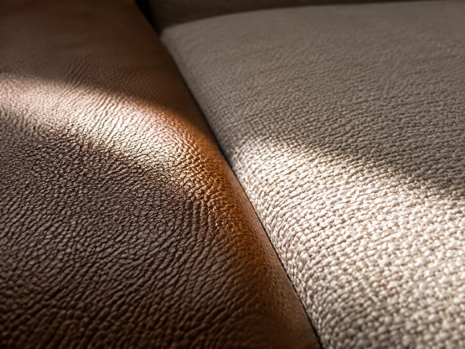 A band of low raking daylight crossing a mid-brown leather seat surface and a pale woven one, the leather returning a soft sheen while the weave stays matte