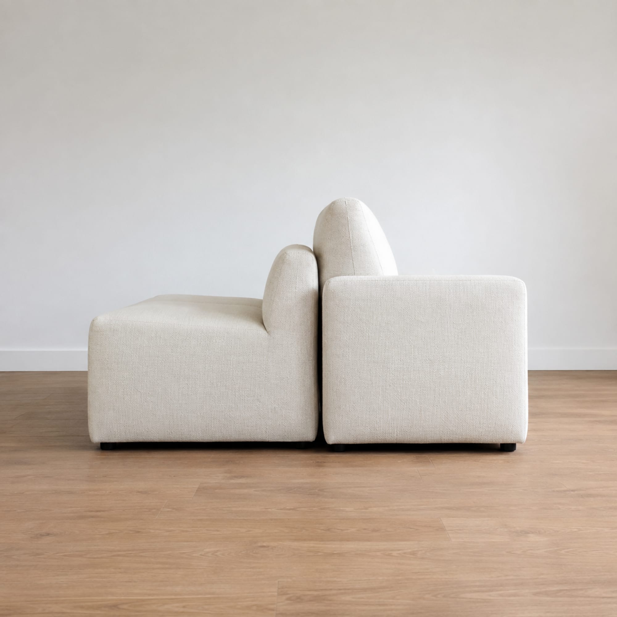 Two sofas in the same pale upholstery standing back to back in an empty room seen from the side, the near one low and deep and the far one taller and more upright