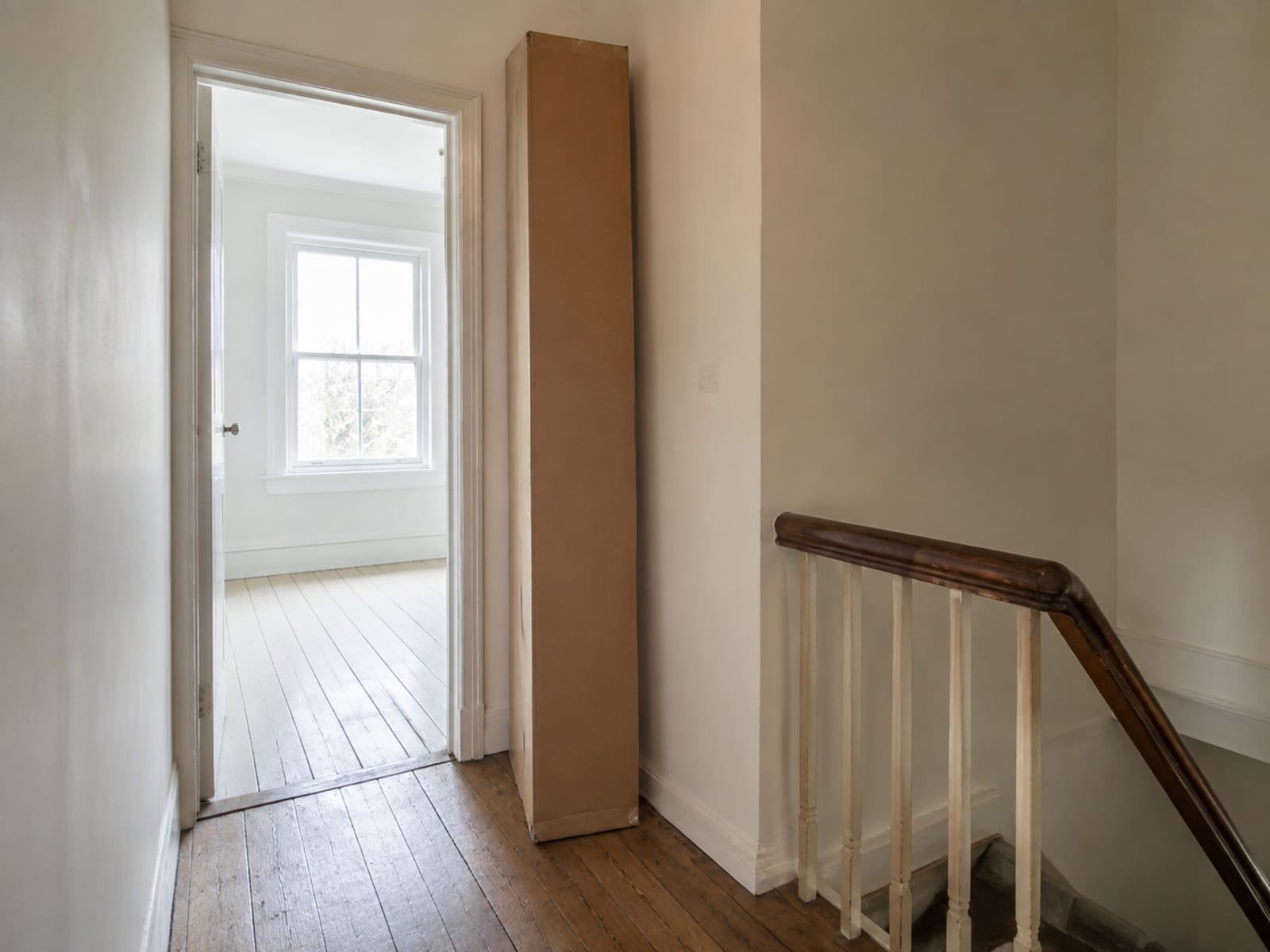 A long flat furniture carton standing on end on an upstairs landing, taller than the bedroom doorway beside it, with the top of the stairs in frame