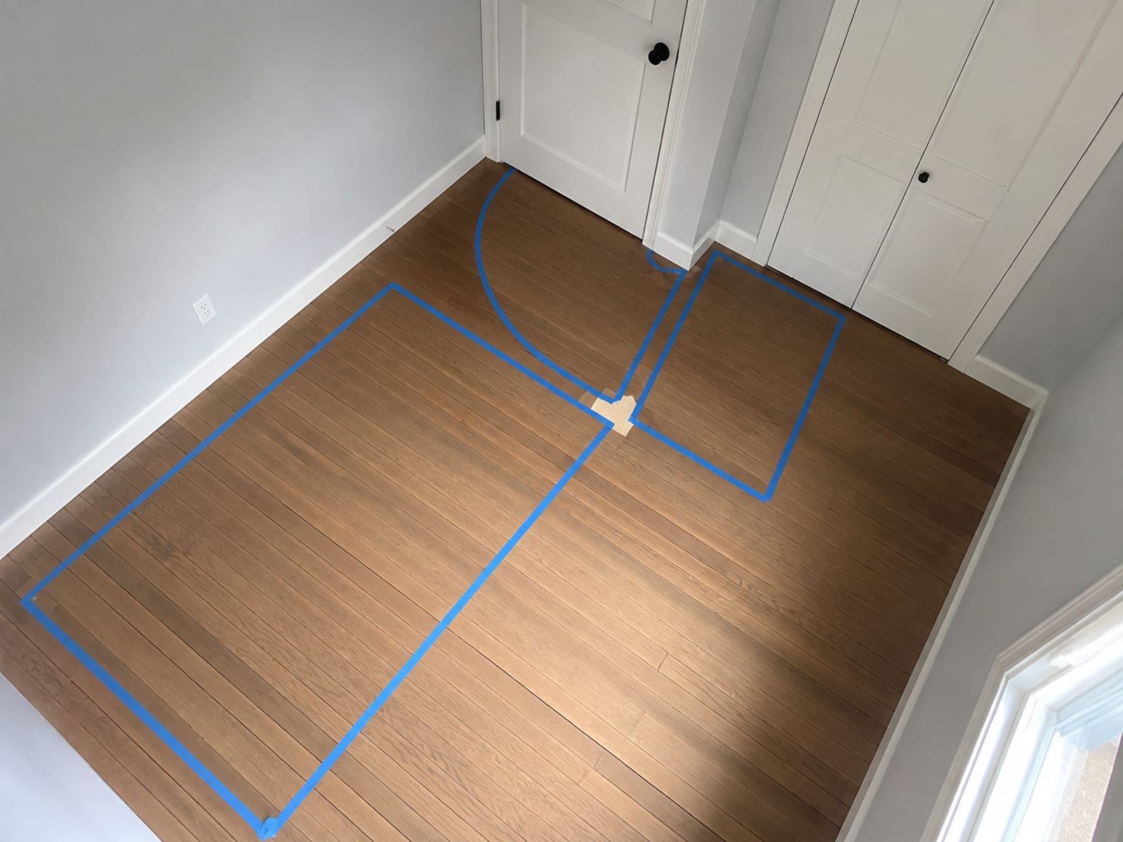 A bare bedroom floor with the bed rectangle, the door's swing arc and the closet's clearance strip all marked out in painters tape
