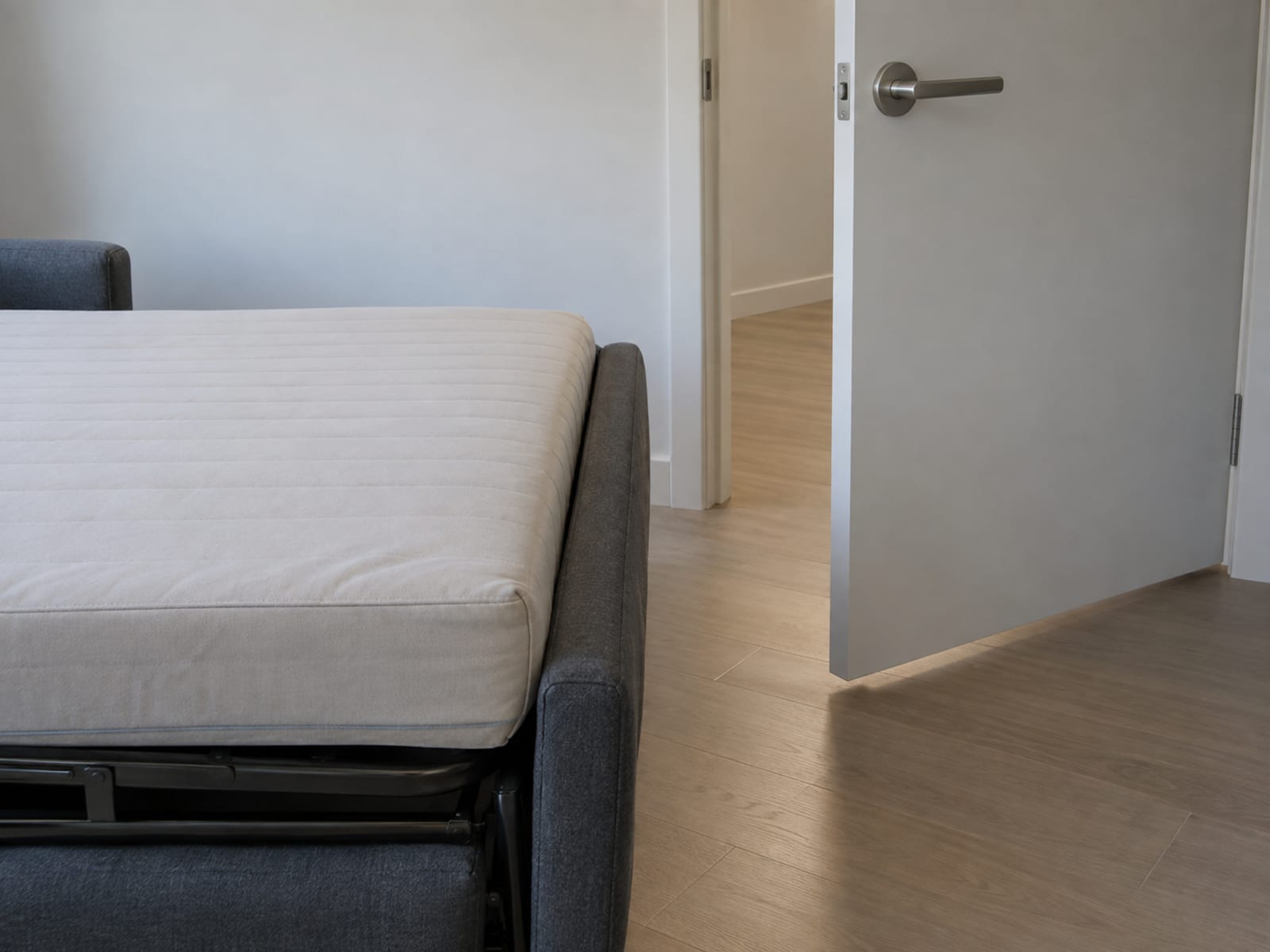 The foot of a fully opened sofa bed in a small daylit bedroom with the room's plain interior door standing open just clear of the mattress's end, the door's lower edge passing a short gap above the floor and a hand's width from the foot of the bed
