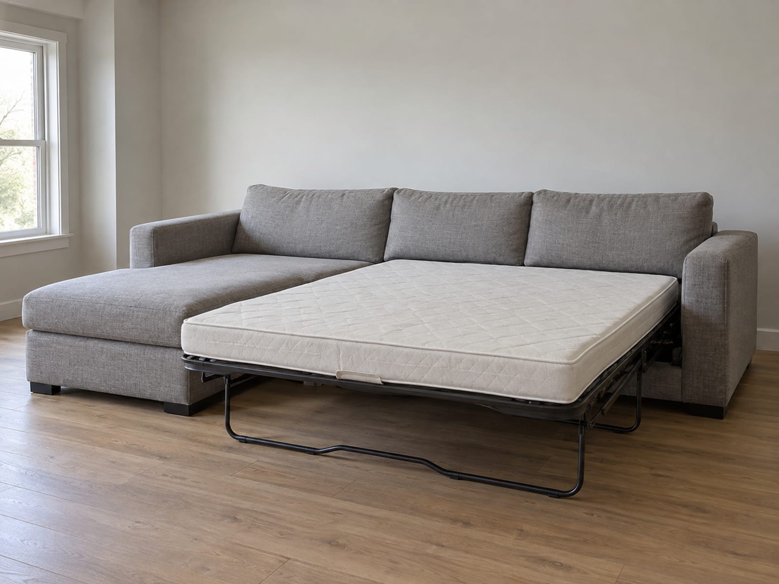 An L-shaped sleeper sectional in a plain daylit room with a pull-out bed drawn fully out from its straight run while its chaise stays in place beside it, photographed from standing height at a three-quarter angle from the open side