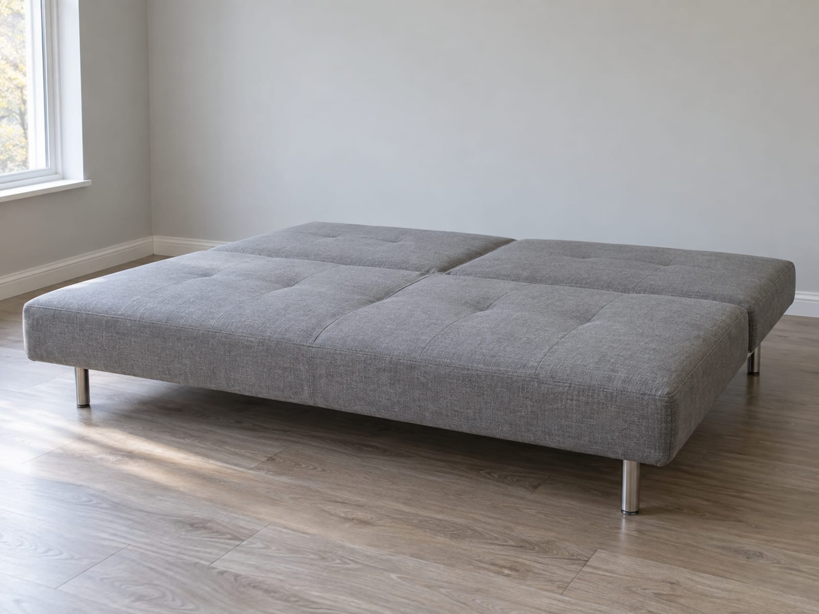 An armless upholstered click-clack style sofa with its back folded fully down into one flat sleeping surface level with the seat, standing on slim metal legs in an empty daylit room, photographed from a low front angle at seat height