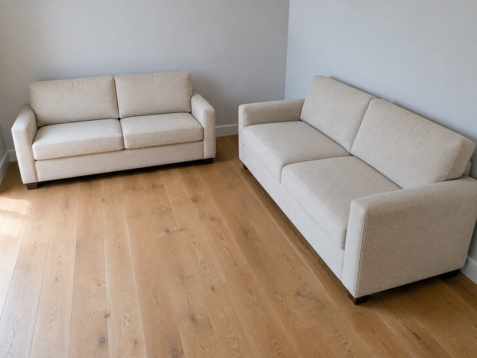 Two sofas of the same length but visibly different depth set at right angles in the corner of a bare room, seen from above the corner