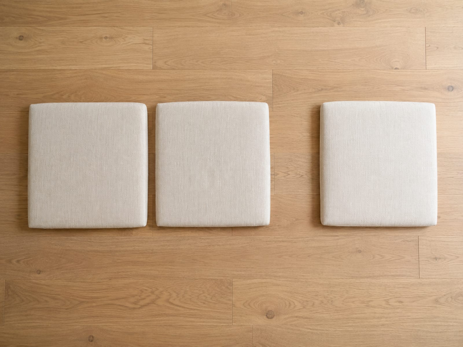 Three identical seat cushions lying flat in a row on bare oak floorboards seen from directly above, with a gap of floor between the second and the third