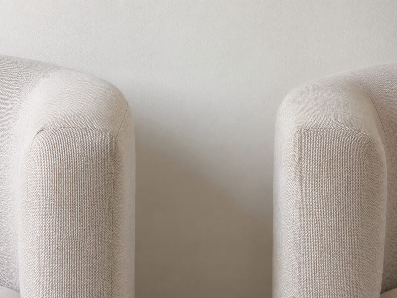 Two sofa arms of identical profile and identical pale fabric seen close and straight on, one at each side of the frame with plain wall between them