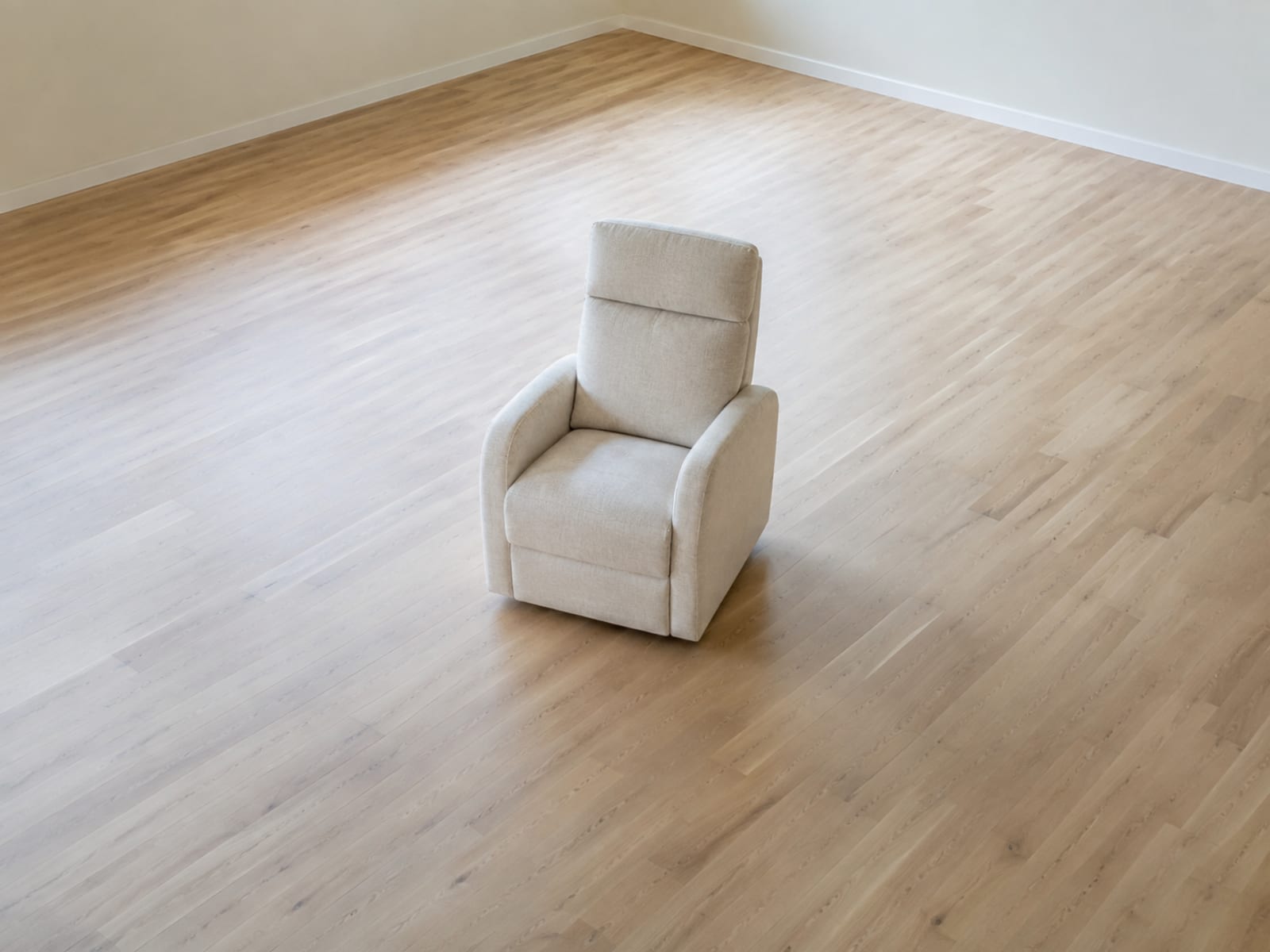 One upholstered armchair standing alone near the middle of an empty room seen from a raised angle, with an unbroken ring of bare floorboards on every side of it