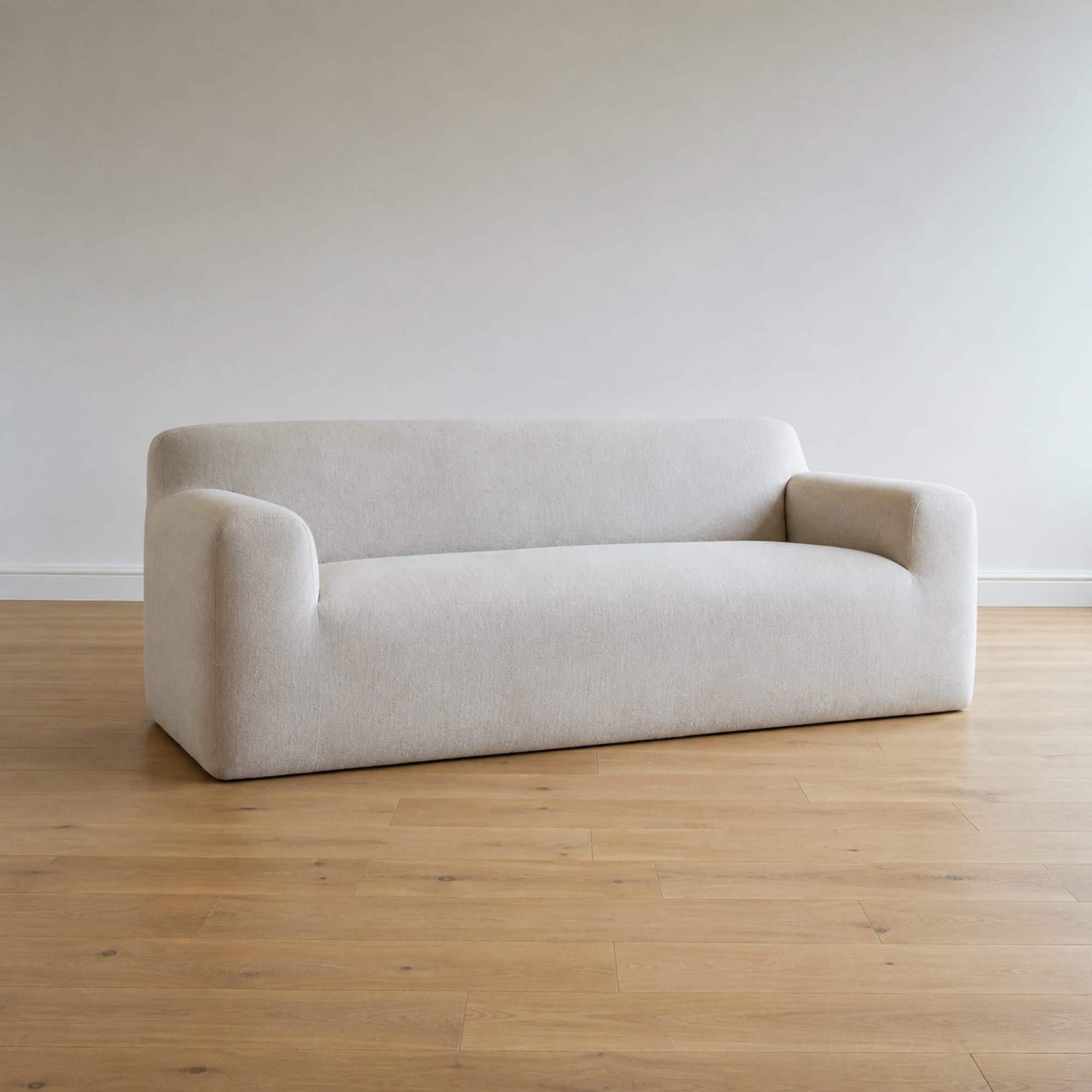A boneless sofa alone in an empty room seen from a low front corner, its base meeting the bare oak floorboards in one unbroken line with no leg or shadow gap