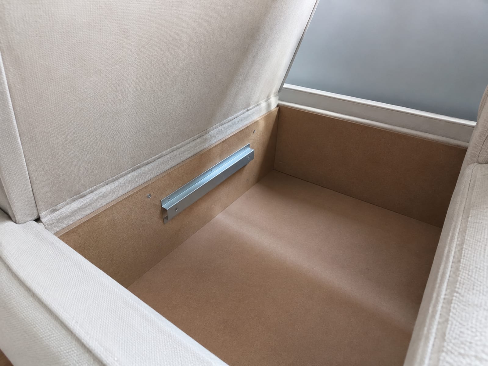 Looking down into an empty under-seat storage well with the upholstered lid propped open and a metal mounting bracket fixed to the inner wall