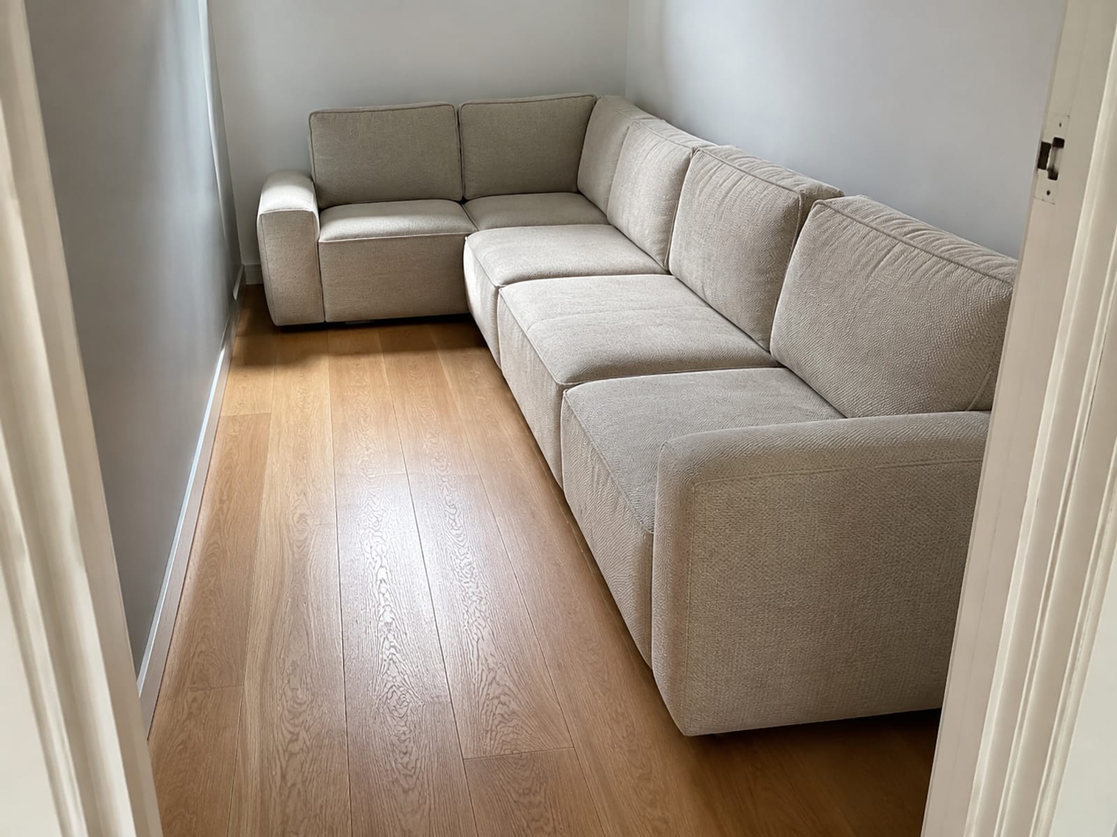 A small living room seen from the doorway, a deep L-shaped modular filling the far corner and a narrow strip of bare floor running past it