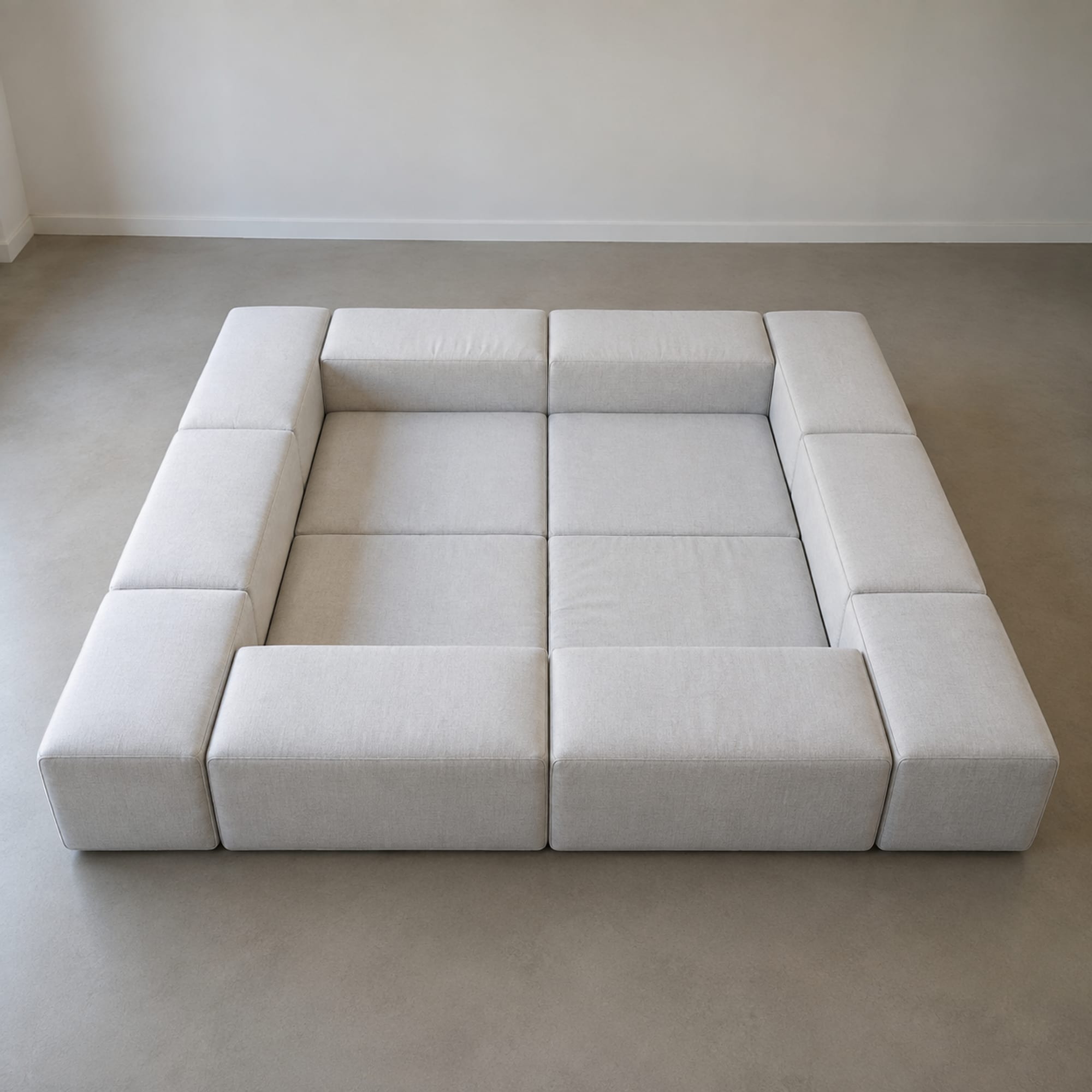 A large sectional arranged into a closed rectangle in a bare room with armless units along the sides and two ottomans filling the middle, seen from above so the whole seating surface reads as one continuous plane