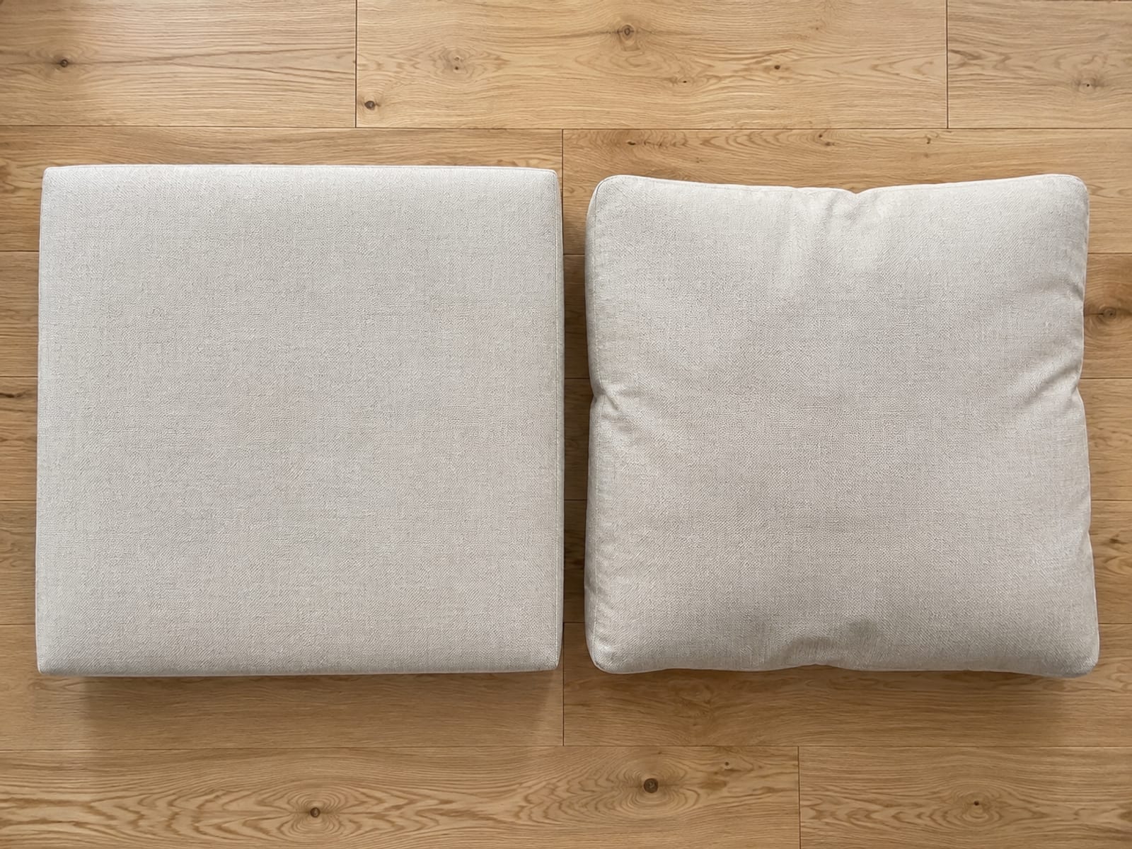 One seat cushion and one back cushion from the same sofa lying side by side on bare oak floorboards seen from directly above, the seat cushion holding a firm square edge and the back cushion slumping at its corners