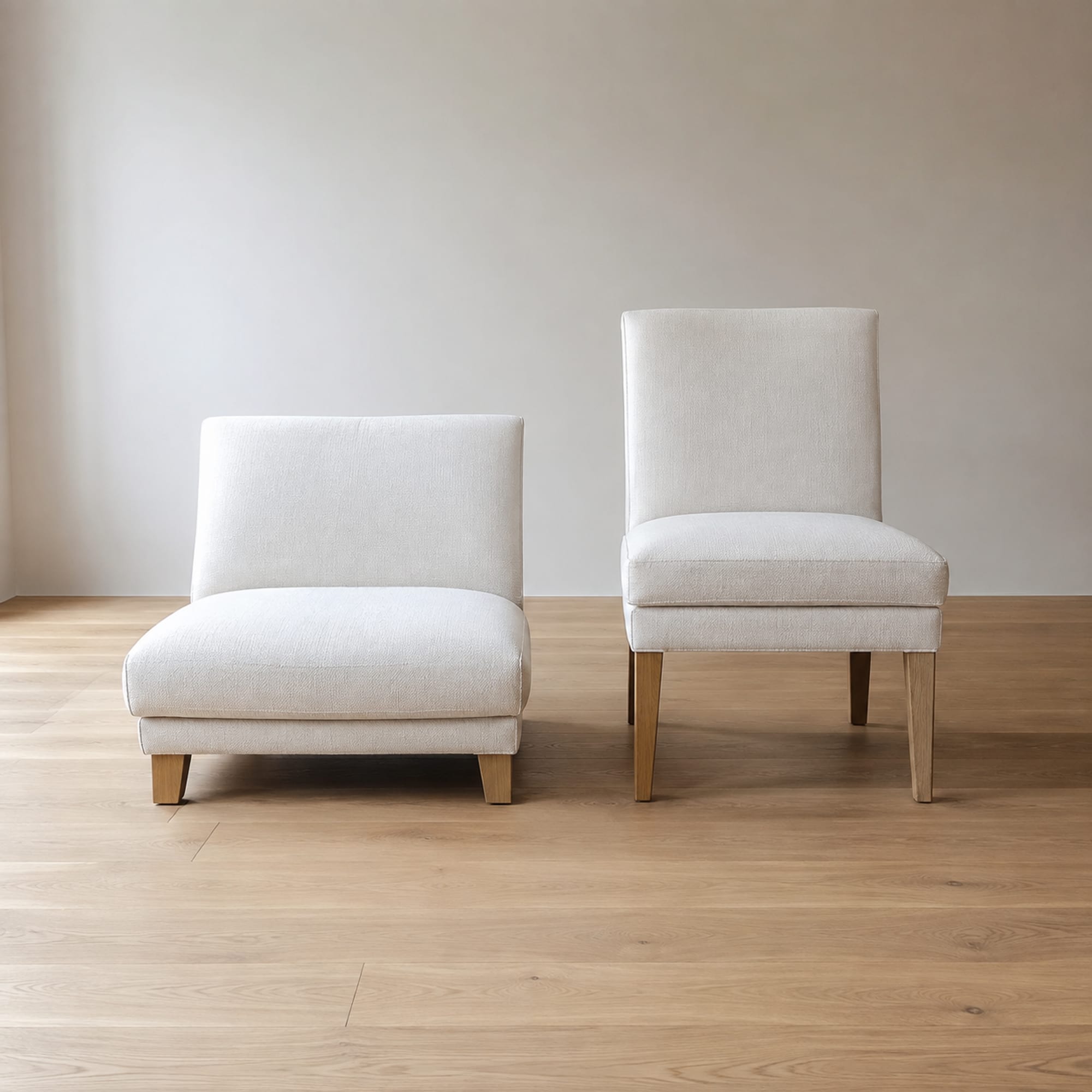 Two upholstered accent chairs of clearly different seat height standing side by side on bare oak floorboards in an empty room, seen square from the front