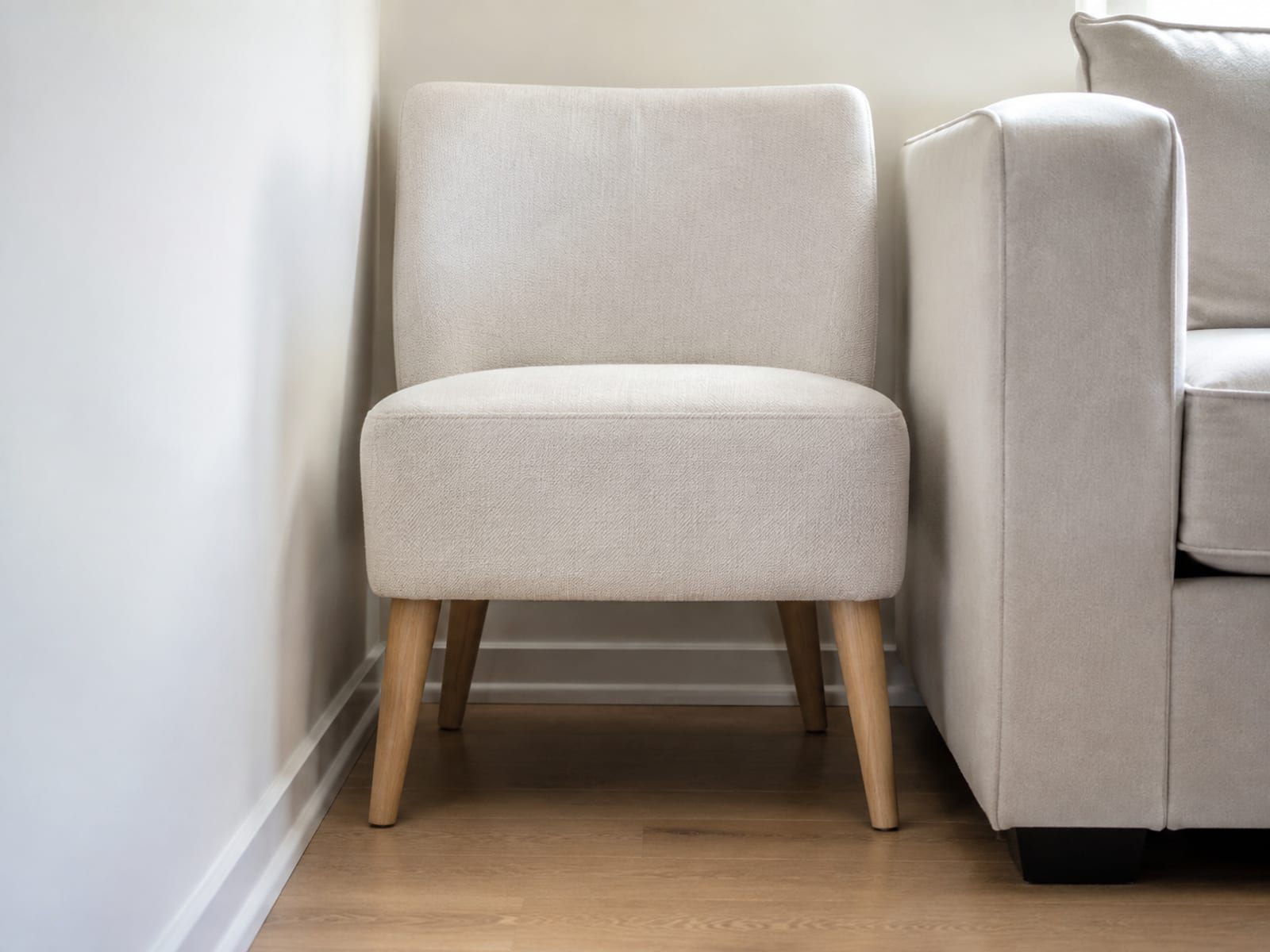 A slim upholstered accent chair standing in the tight space between a plain wall and the end of a sofa arm, with only a small clearance at each side
