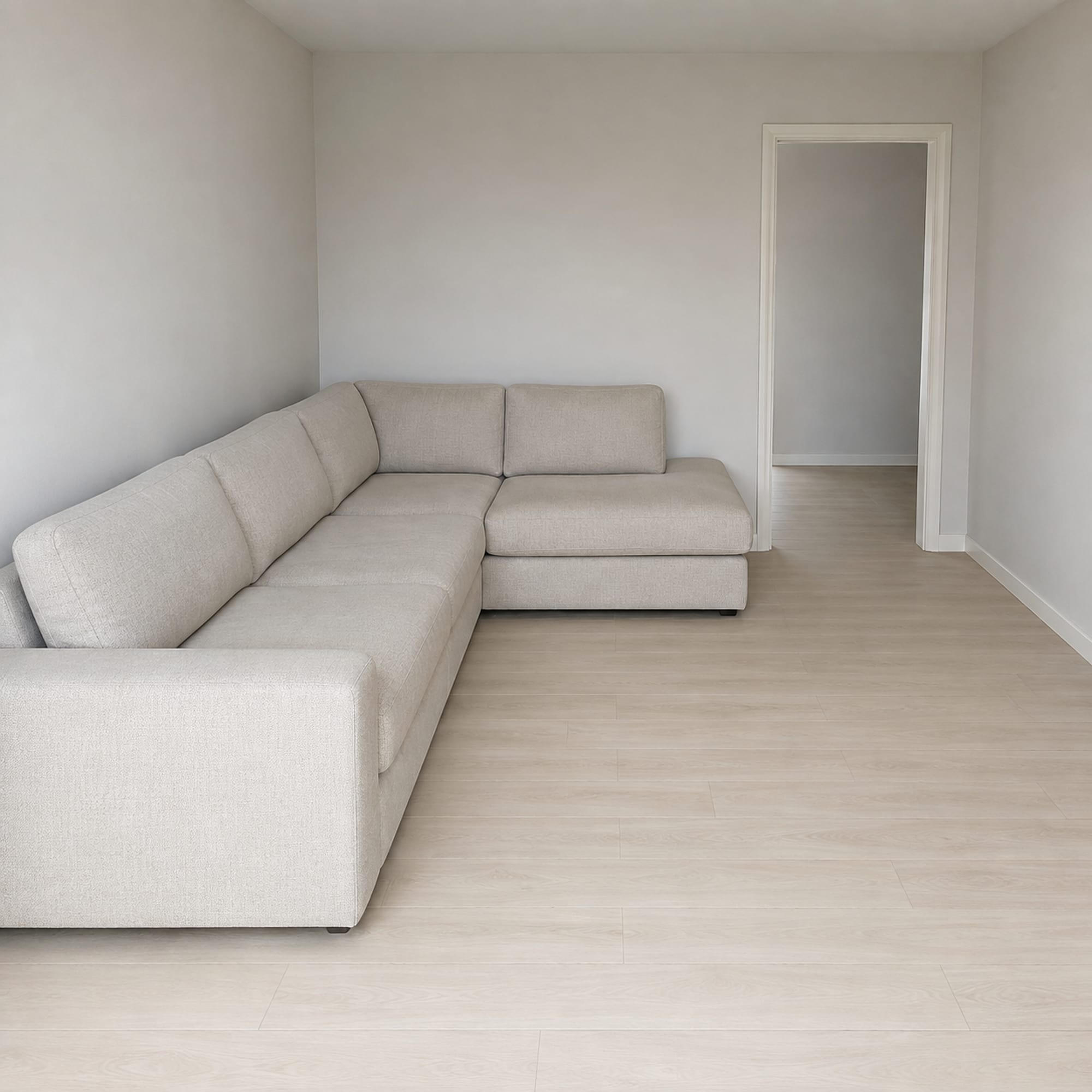 An L-shaped sectional standing against two walls of a small bare room, seen from the opposite corner, with a narrow strip of clear floor running between its front edge and the far wall