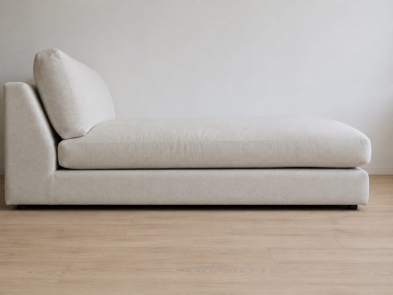 A sectional chaise end seen square from the side at low height against a plain wall, its full front-to-back reach filling the width of the frame with bare floor beyond
